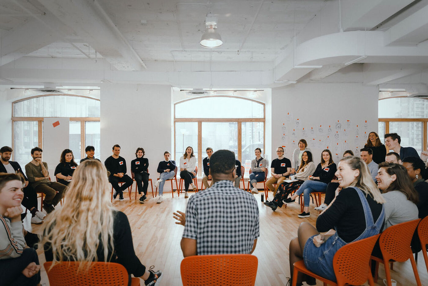 A group of people are sitting in a circle in a bright, open room. They appear to be engaged in a meeting or discussion. Some are seated in chairs, while others are standing or walking around.