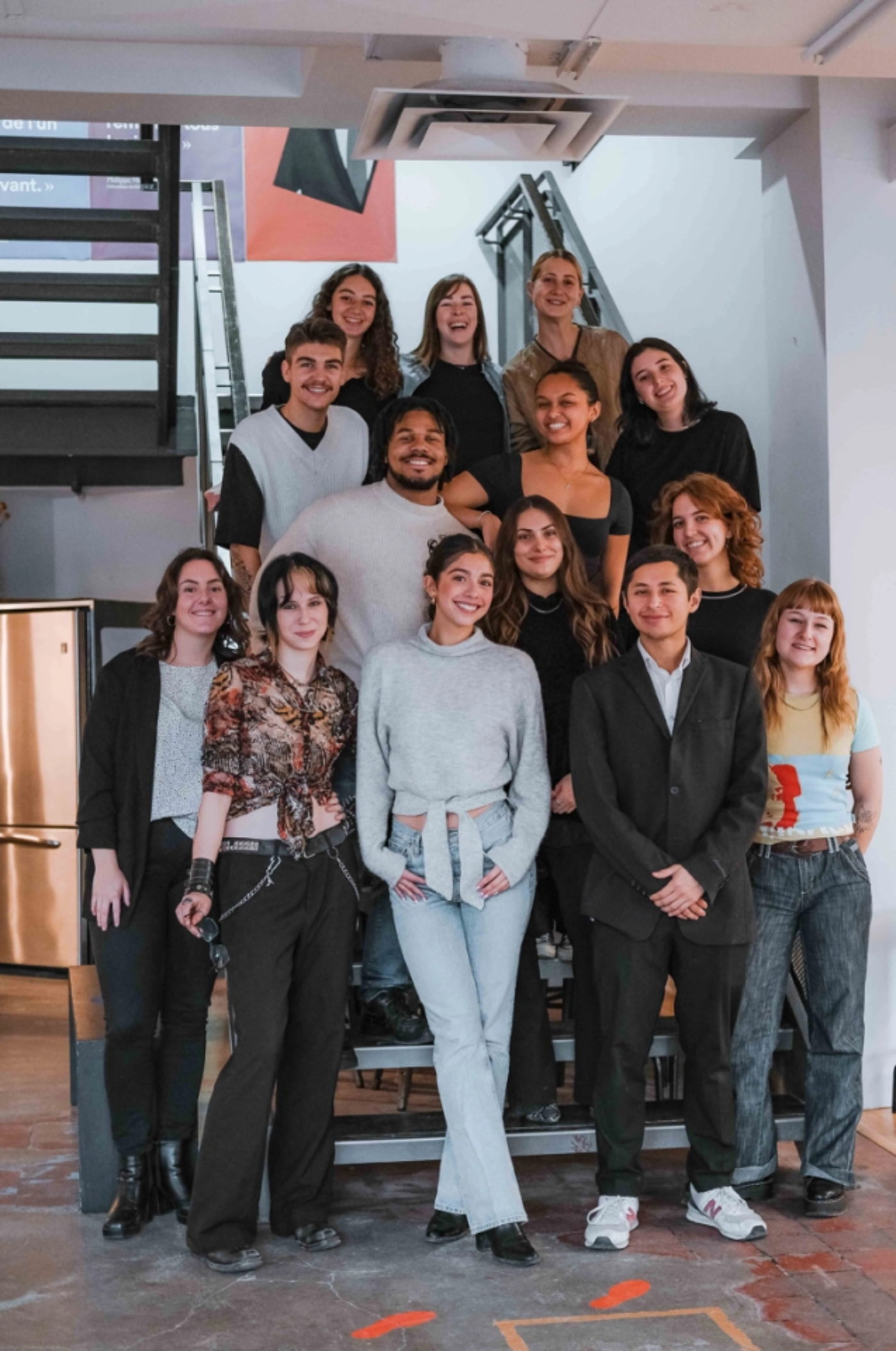 A group of approximately 14 young adults are posing for a photo on a staircase. They are dressed in casual clothing and are smiling. The background is a modern, industrial-style interior with exposed ductwork and a metal staircase.