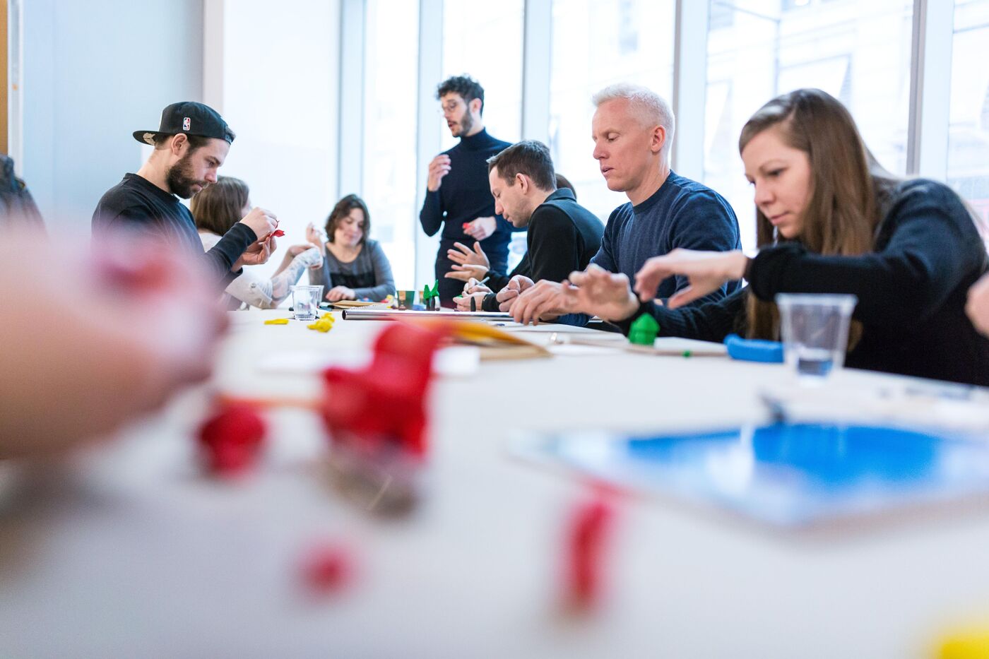 A group of people are gathered around a table, possibly engaged in a collaborative activity or meeting. They appear focused and involved, with various objects scattered on the table, suggesting a hands-on project.
