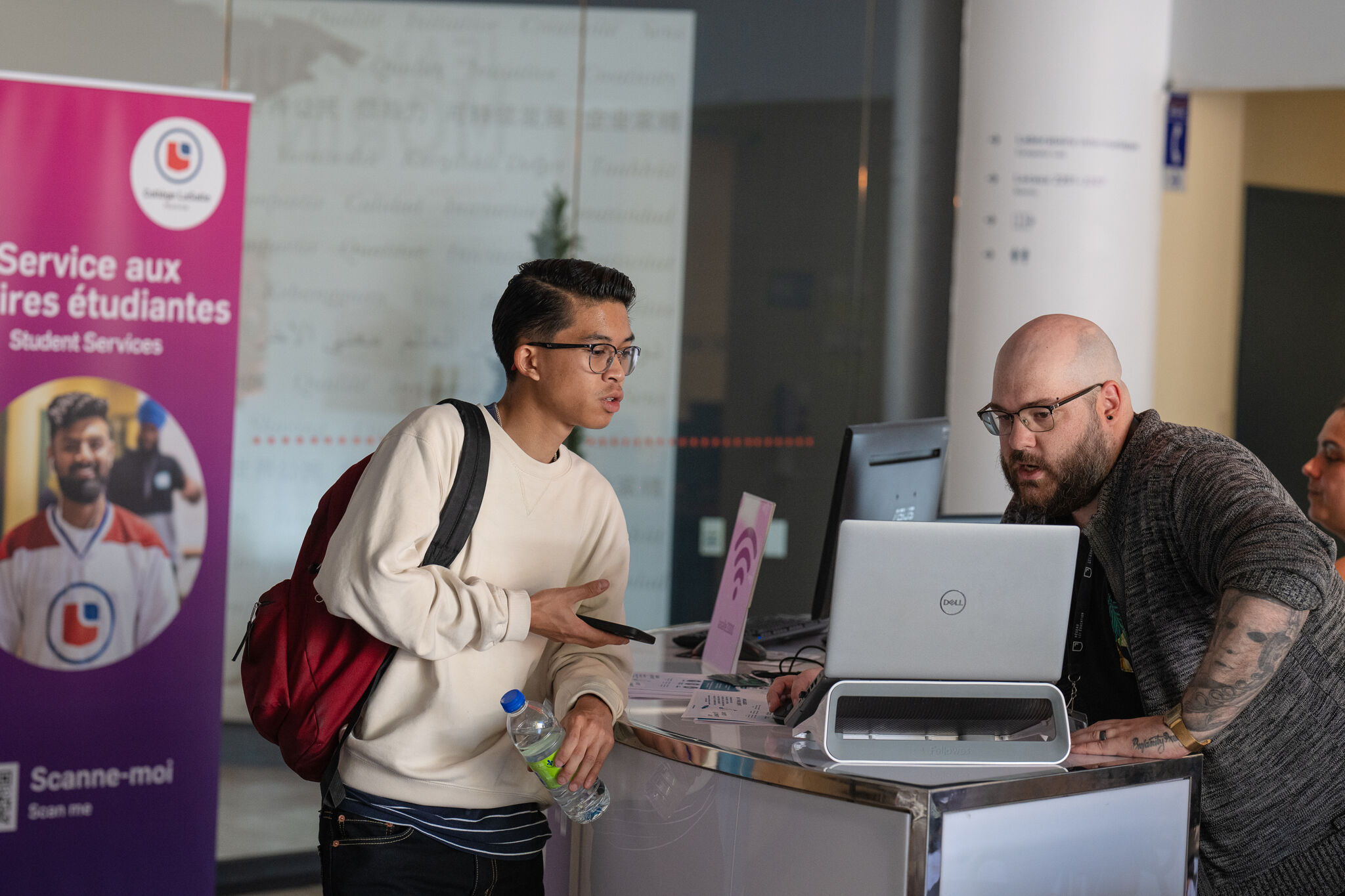 A young student, holding a water bottle and backpack, inquires at a university service desk staffed by an attentive personnel.