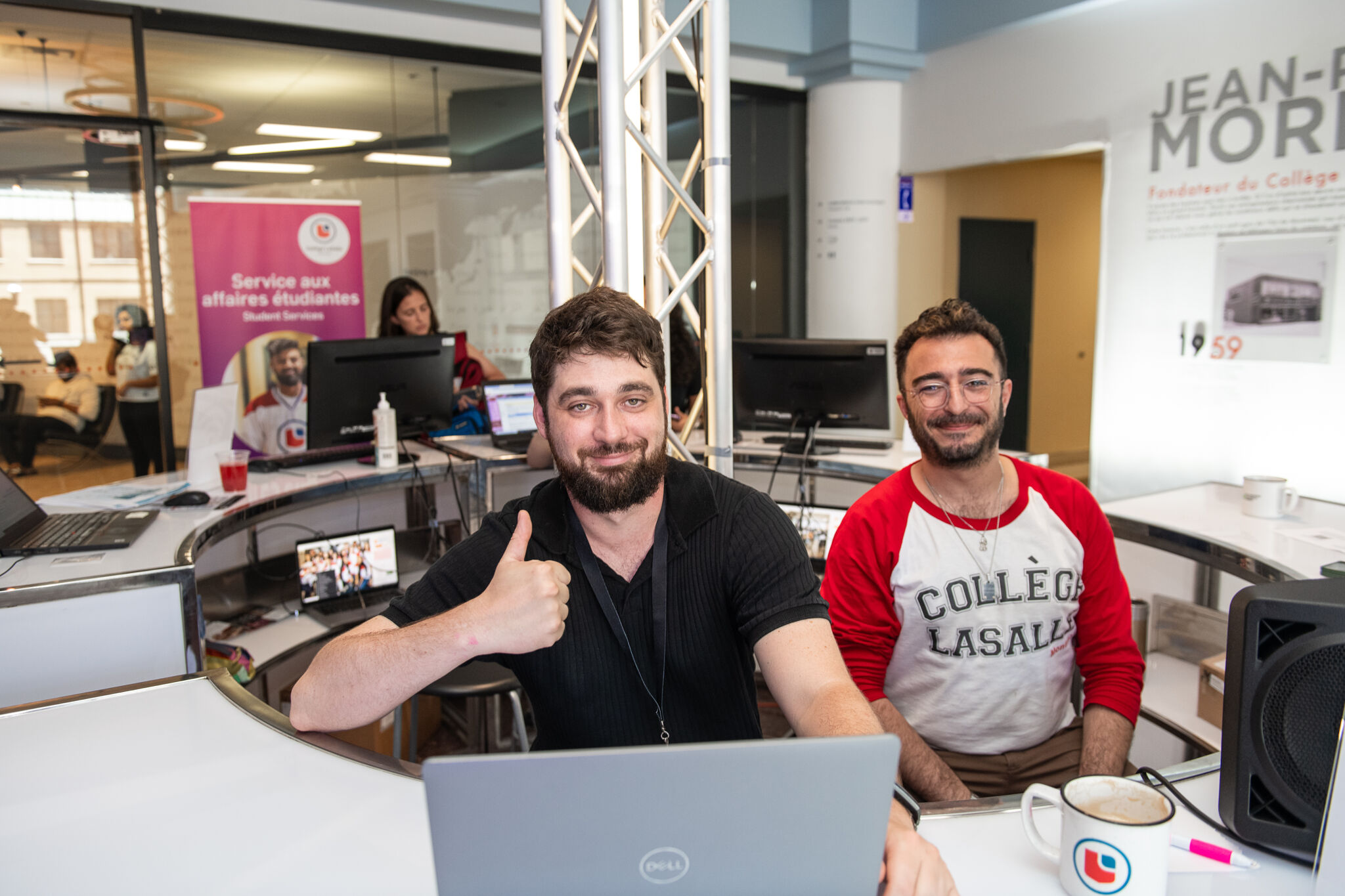 Two men smiling at a desk with laptops in a bustling office environment, one giving a thumbs-up, promoting a collaborative atmosphere.