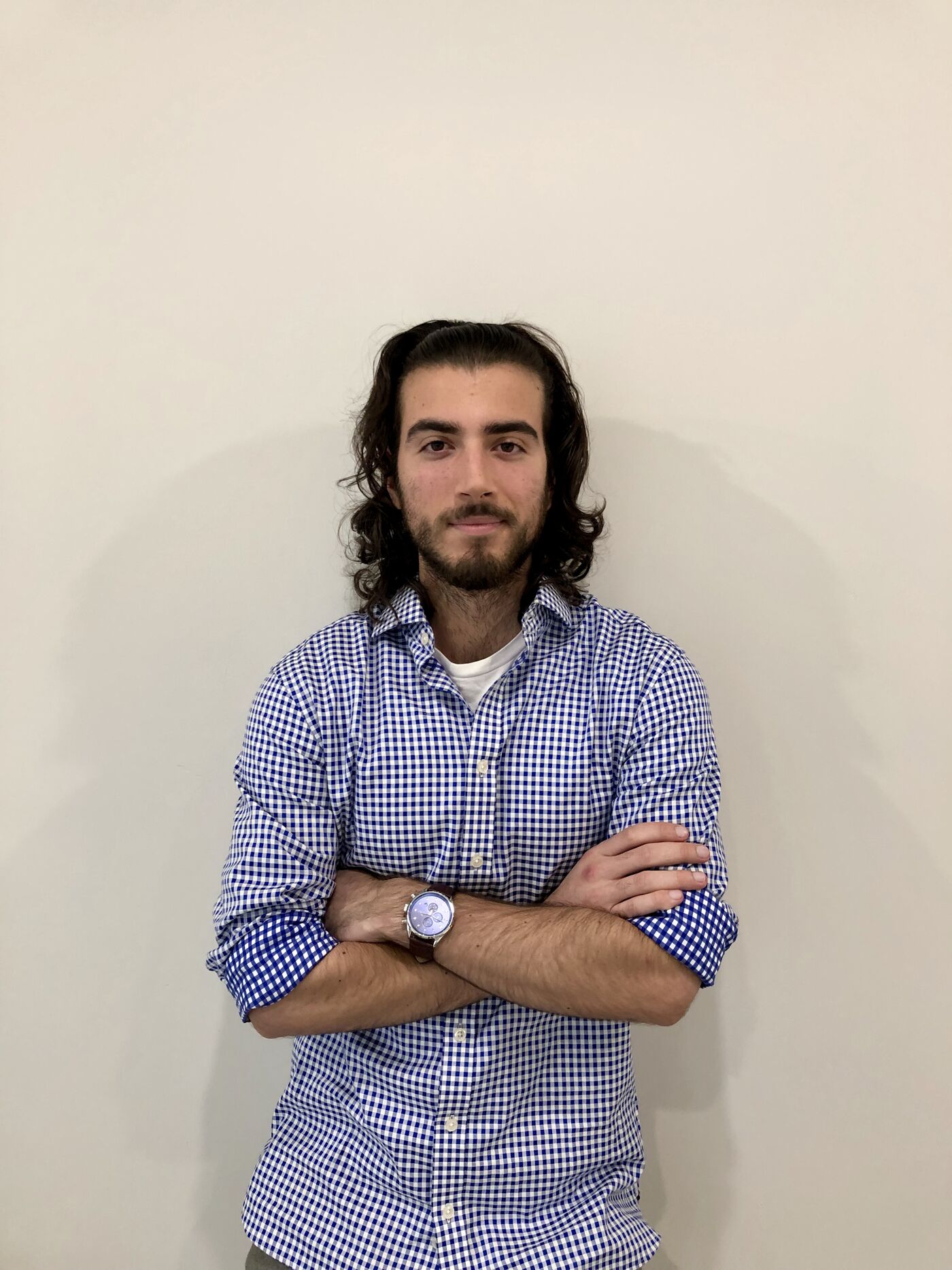 A young man with long curly hair, wearing a blue and white checkered shirt, stands with crossed arms and a watch on his left wrist, against a plain background.