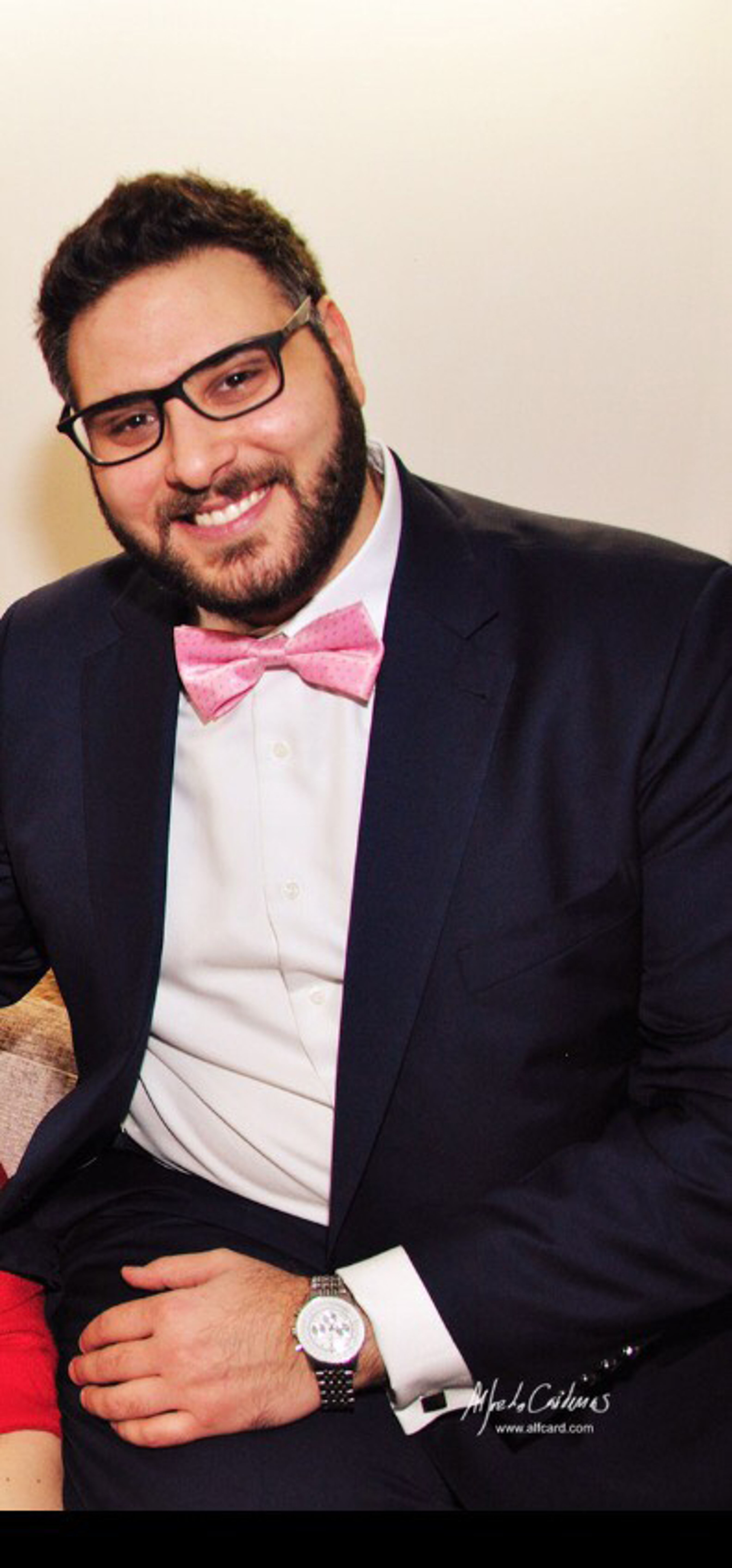 A cheerful man with glasses, wearing a black suit, white shirt, and a pink bow tie, sits with a watch on his wrist.