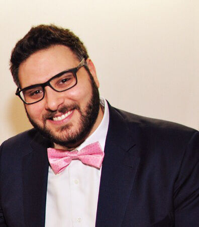 A cheerful man with glasses, wearing a black suit, white shirt, and a pink bow tie, sits with a watch on his wrist.