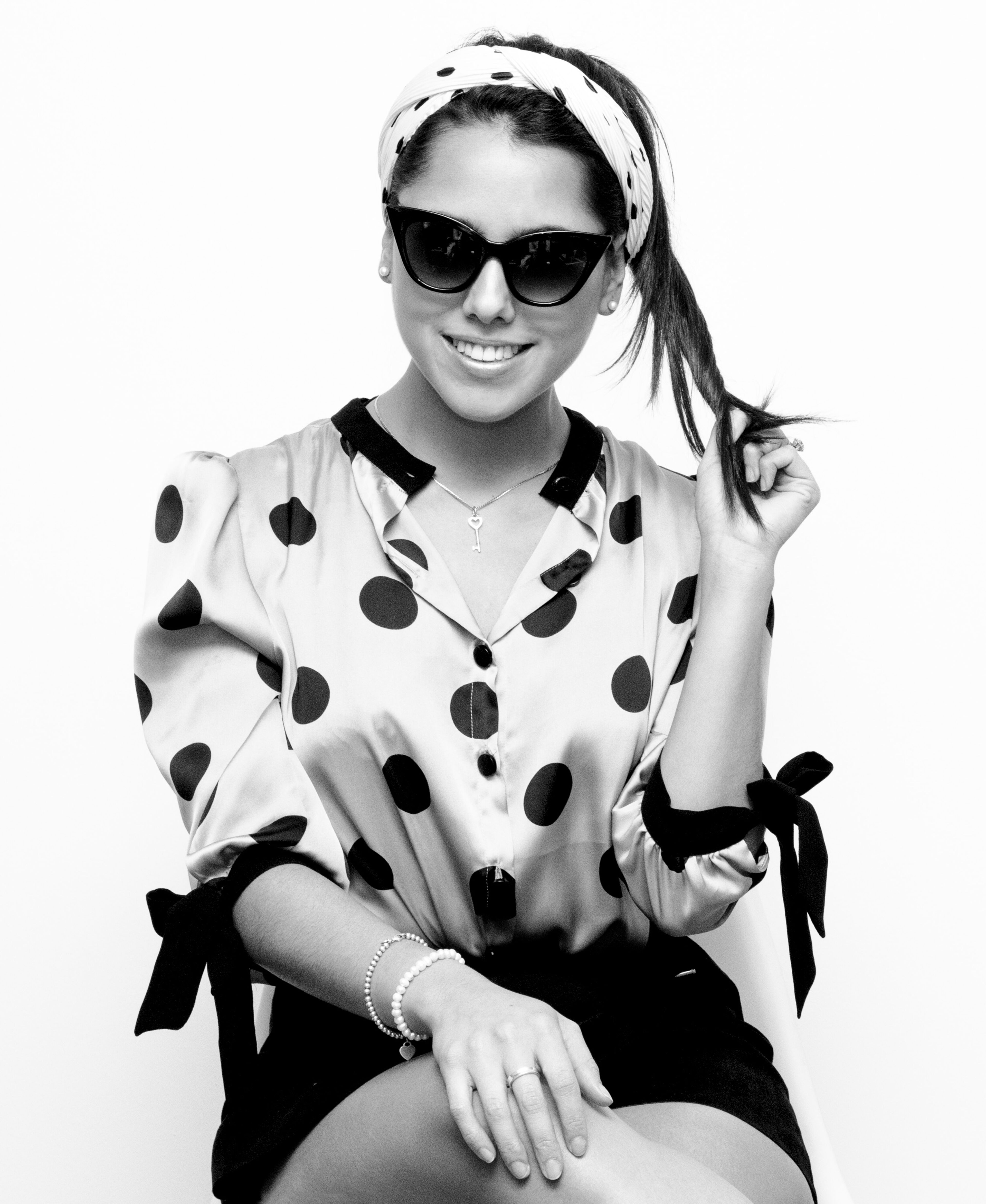 A cheerful woman in a polka-dotted blouse, accessorized with sunglasses, a headscarf, and jewelry, poses playfully in a black and white photo.