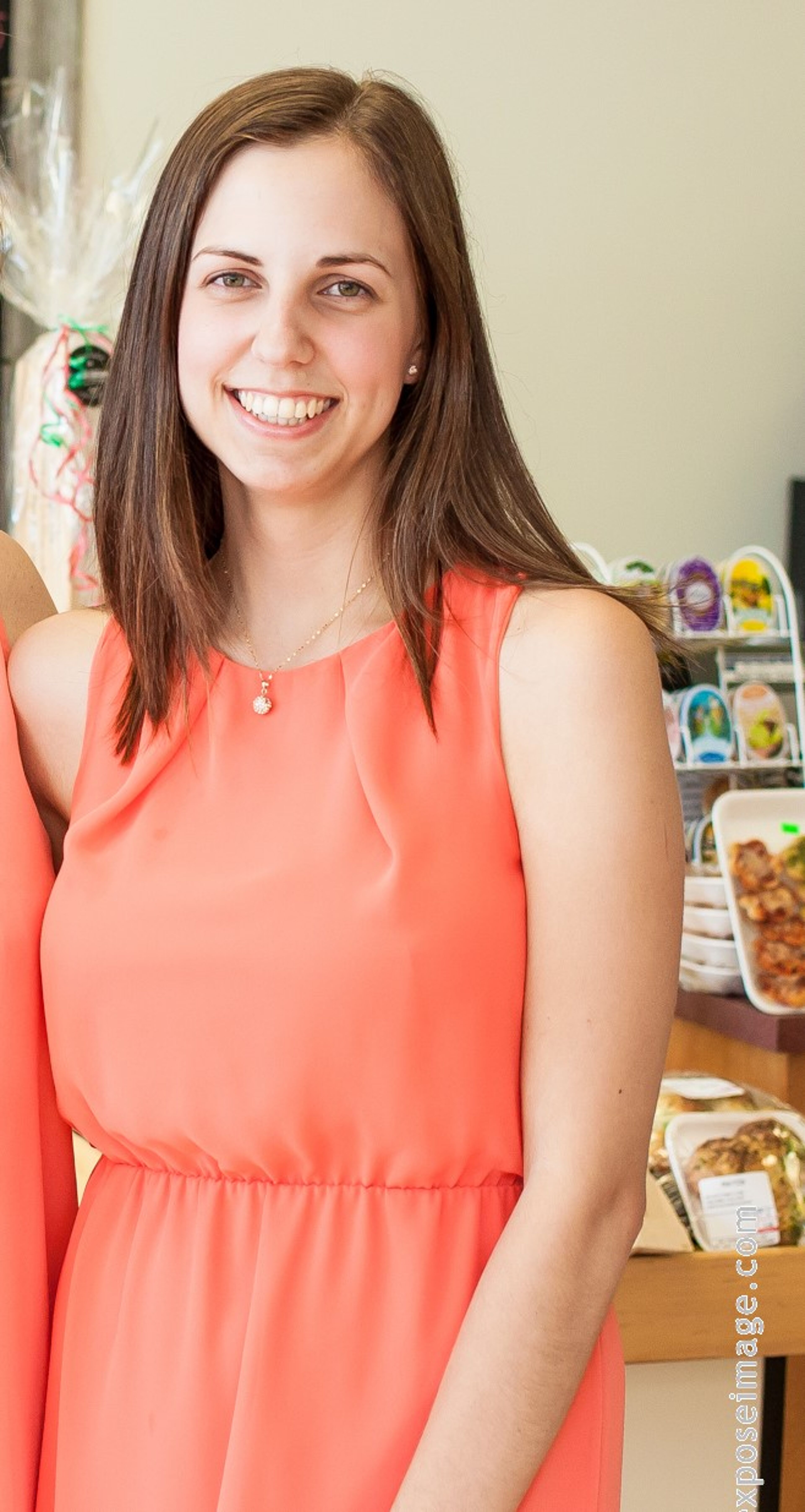 A cheerful young woman in a sleeveless coral dress with a delicate necklace stands in a room with a buffet in the background.