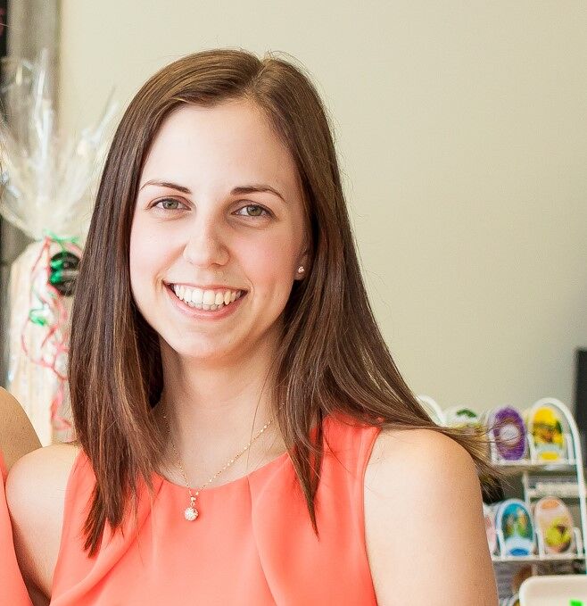 A cheerful young woman in a sleeveless coral dress with a delicate necklace stands in a room with a buffet in the background.