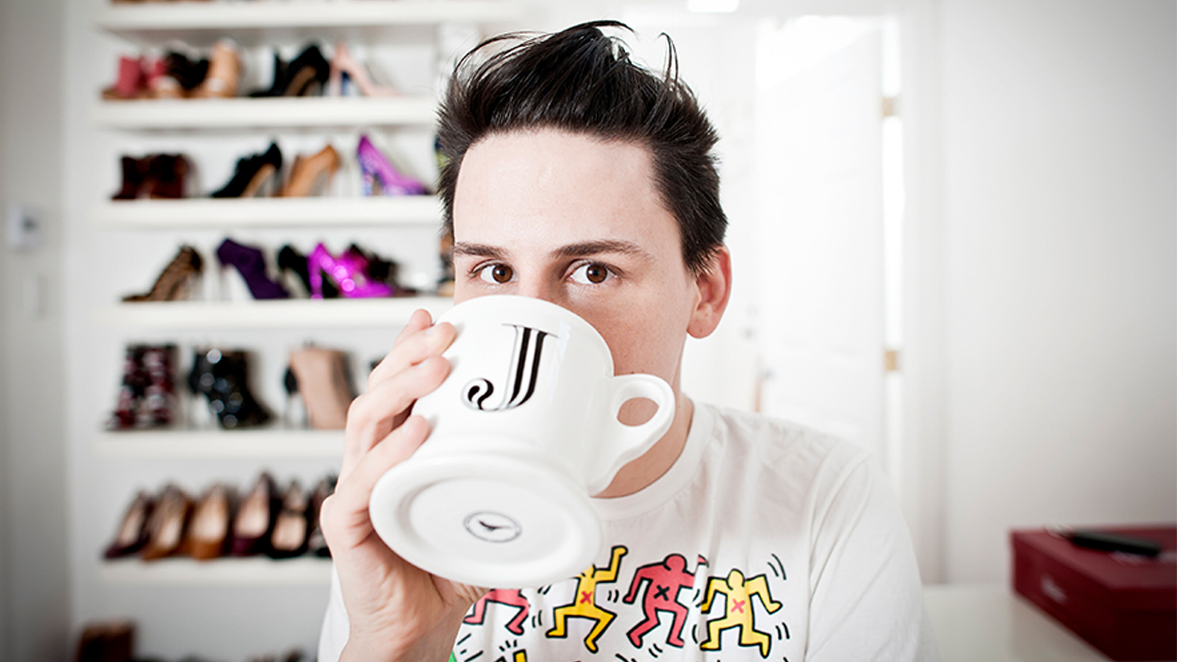 A young man with an edgy hairstyle sips from a creatively designed coffee mug, his gaze meeting the camera, with a backdrop of a colorful shoe collection.