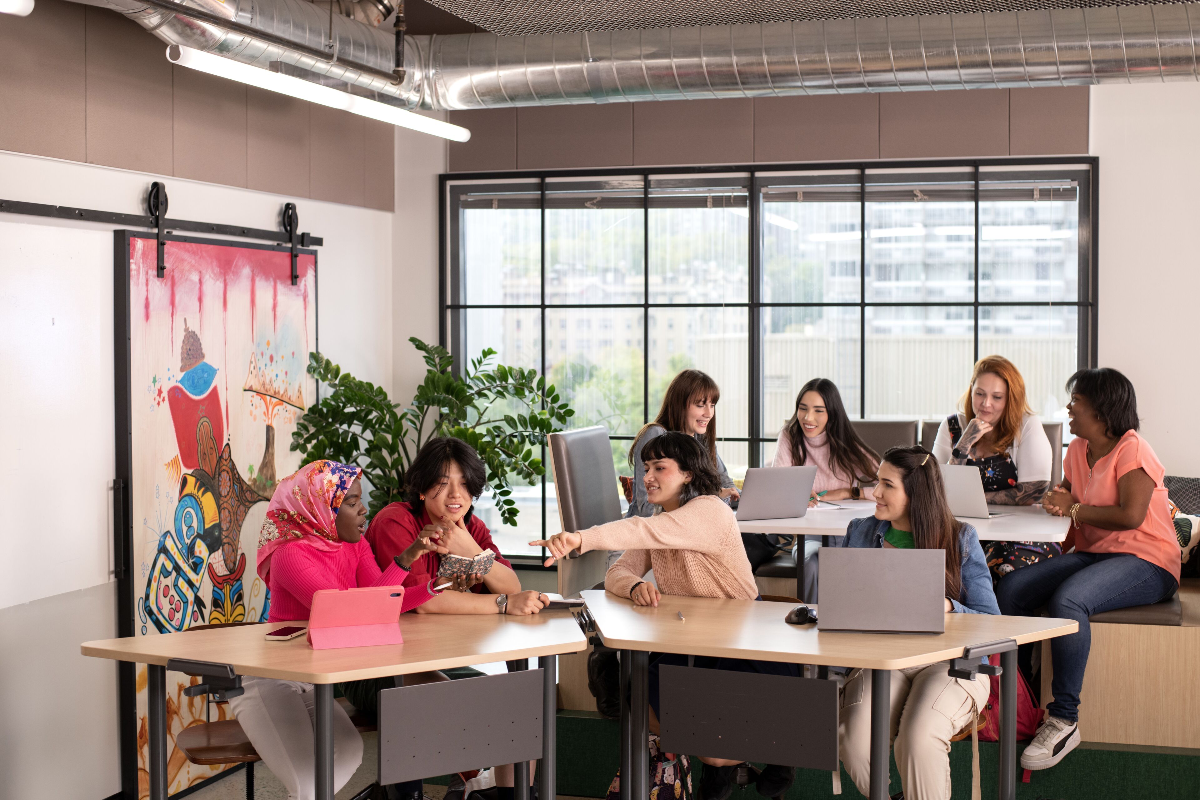 A group of eight diverse women engaging in a lively discussion around a table with laptops in a well-lit office space, adorned with modern art and plants.