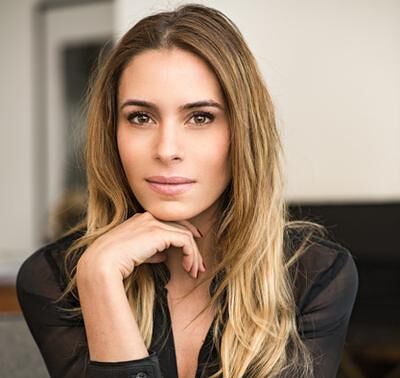 A poised woman with long blonde hair, resting her chin on her hand, wearing a sophisticated black outfit.