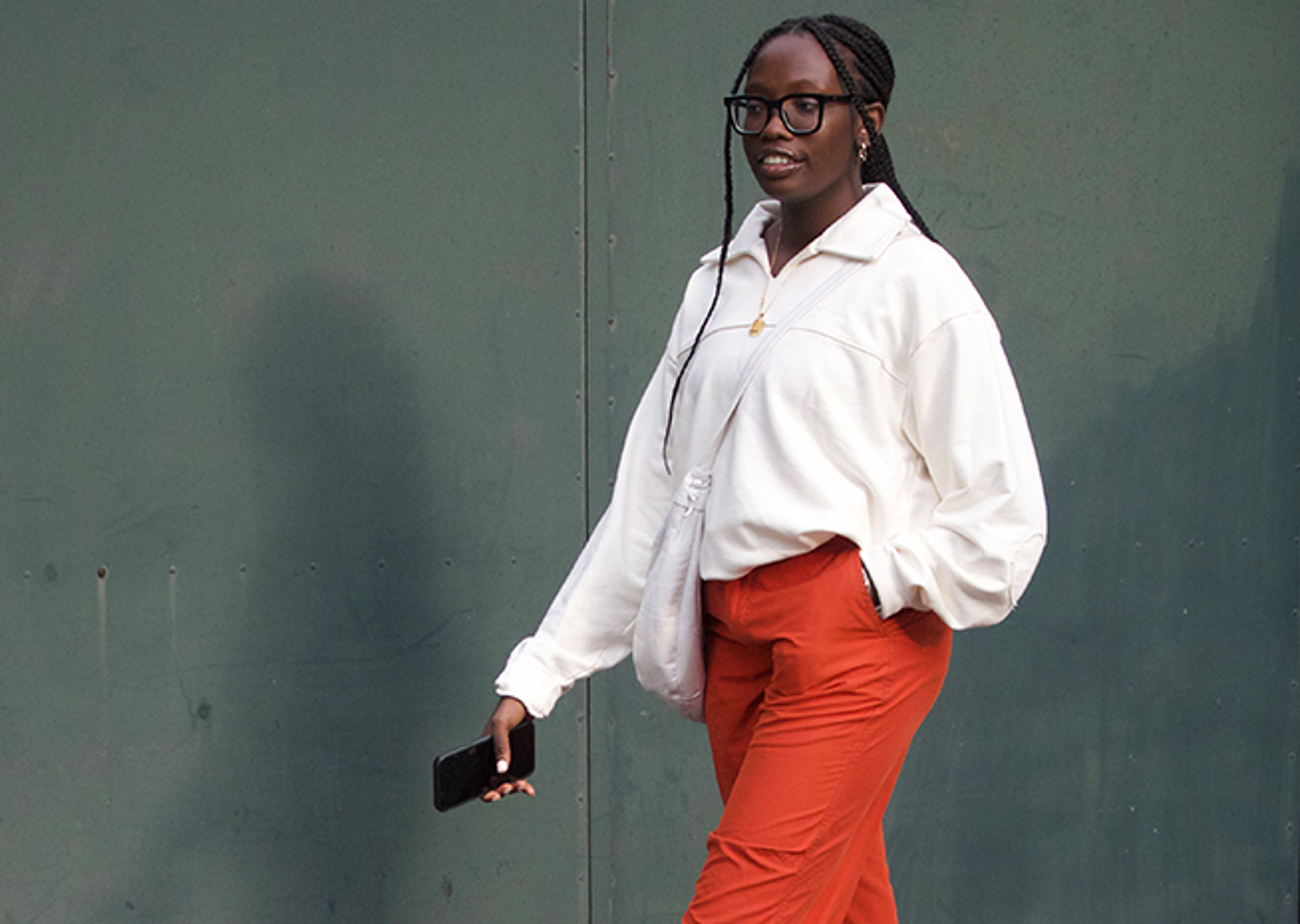 A woman in a white shirt and burnt orange trousers walks confidently, phone in hand, against a green backdrop.