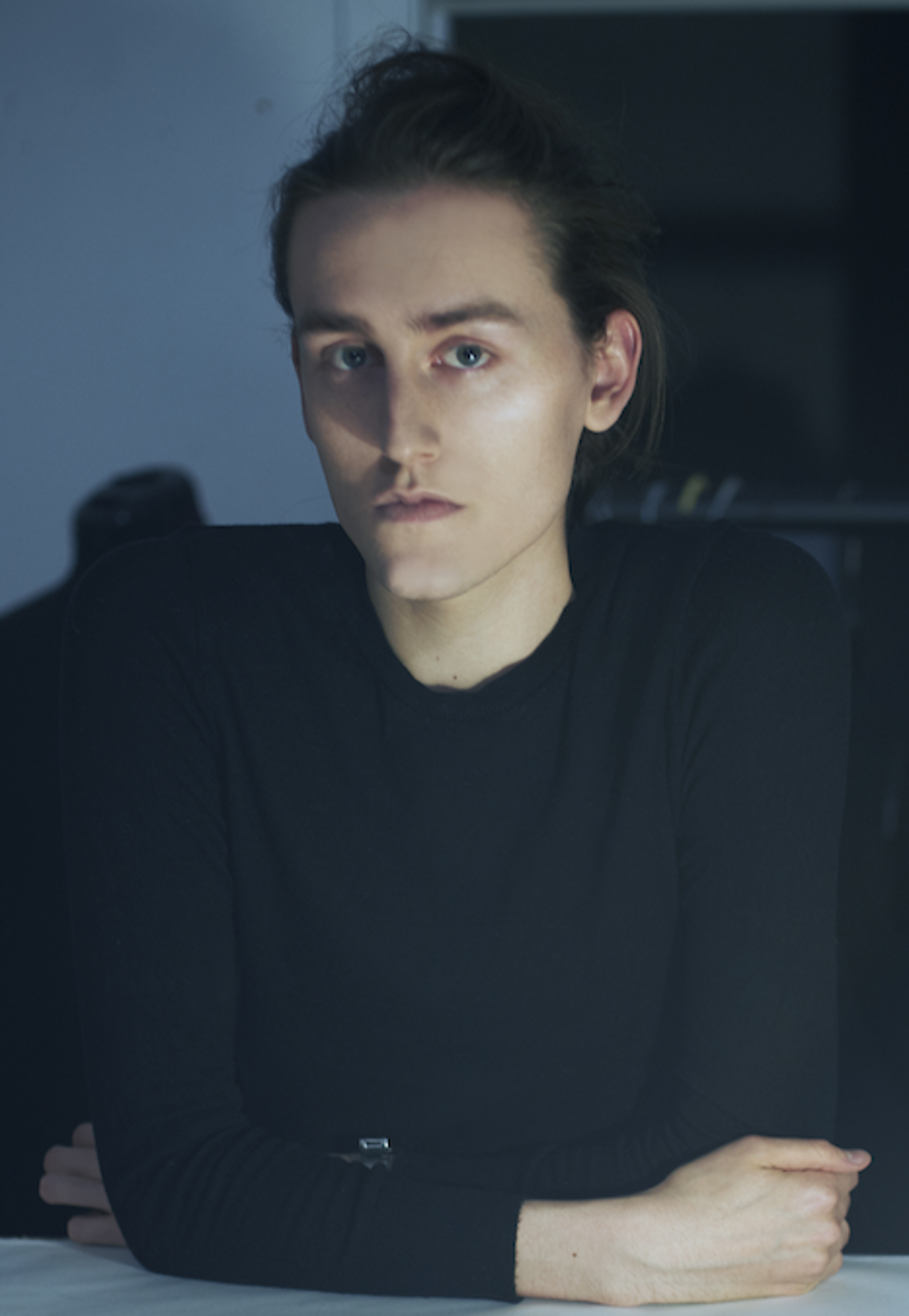 A young adult with a thoughtful expression, lit by soft, cool light, wearing a black shirt.