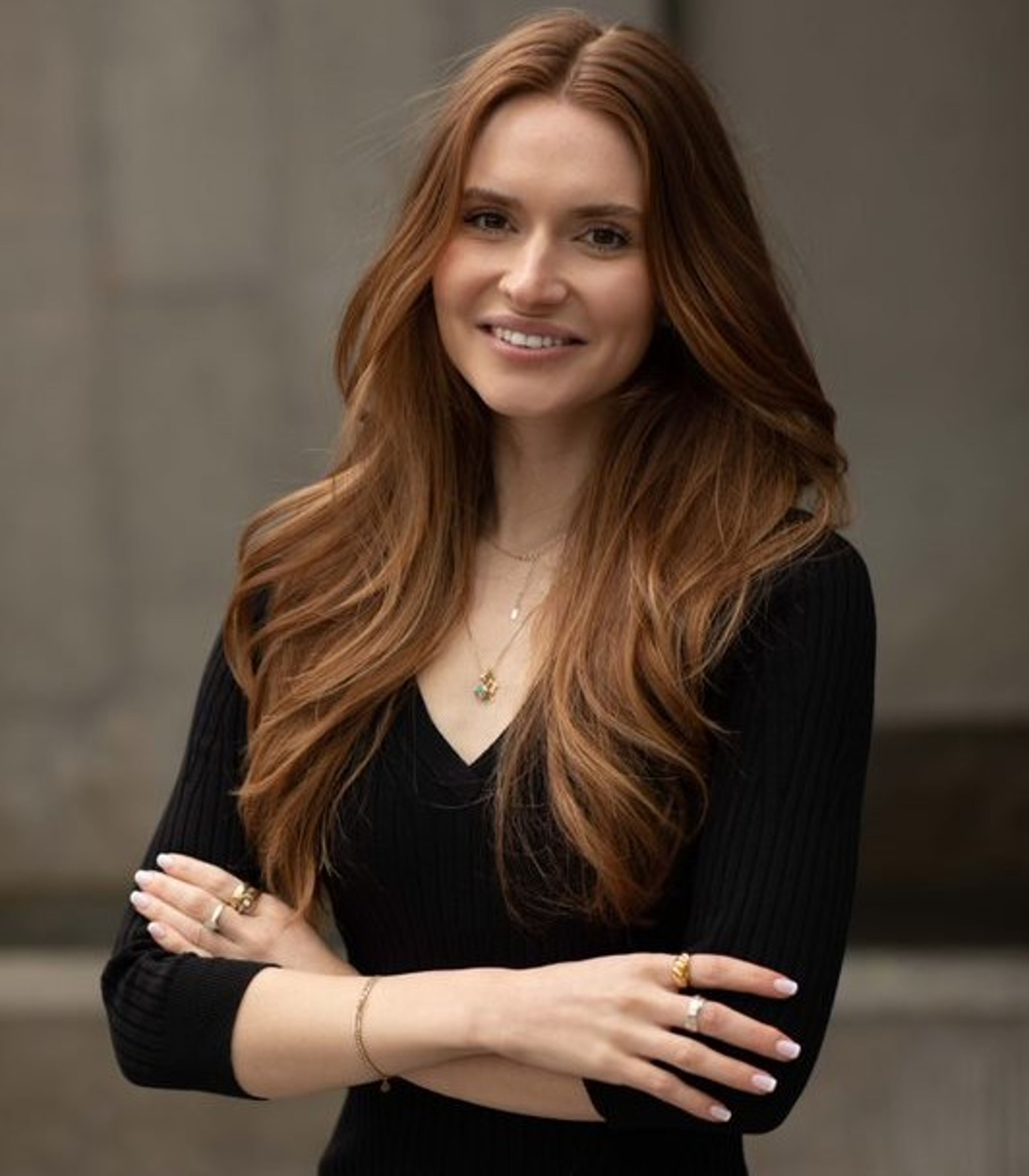 A woman with long auburn hair smiles gently, wearing a black V-neck top and gold jewelry.