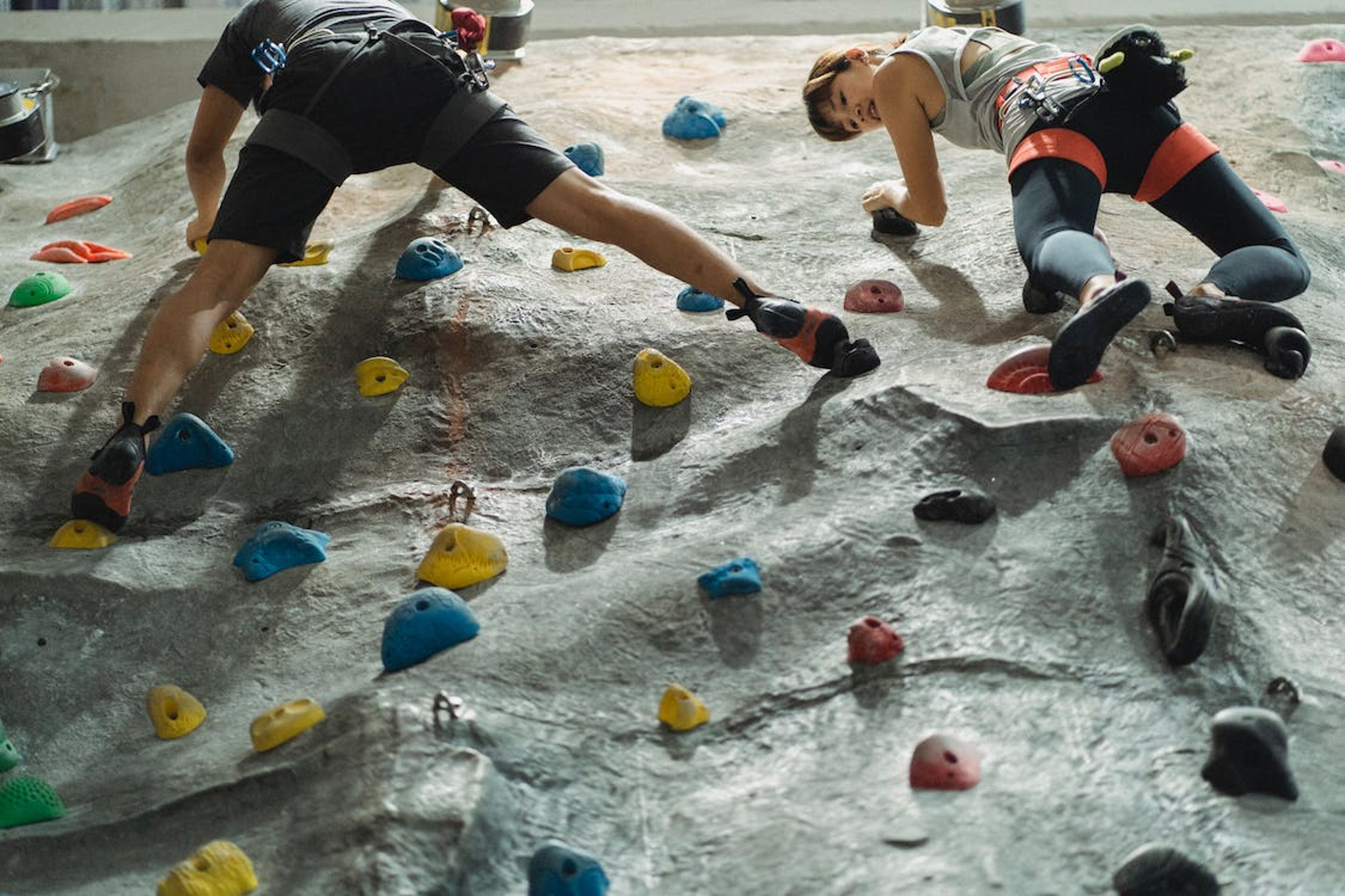 Two climbers practice on an artificial rock wall, using various holds to ascend, secured with harnesses.