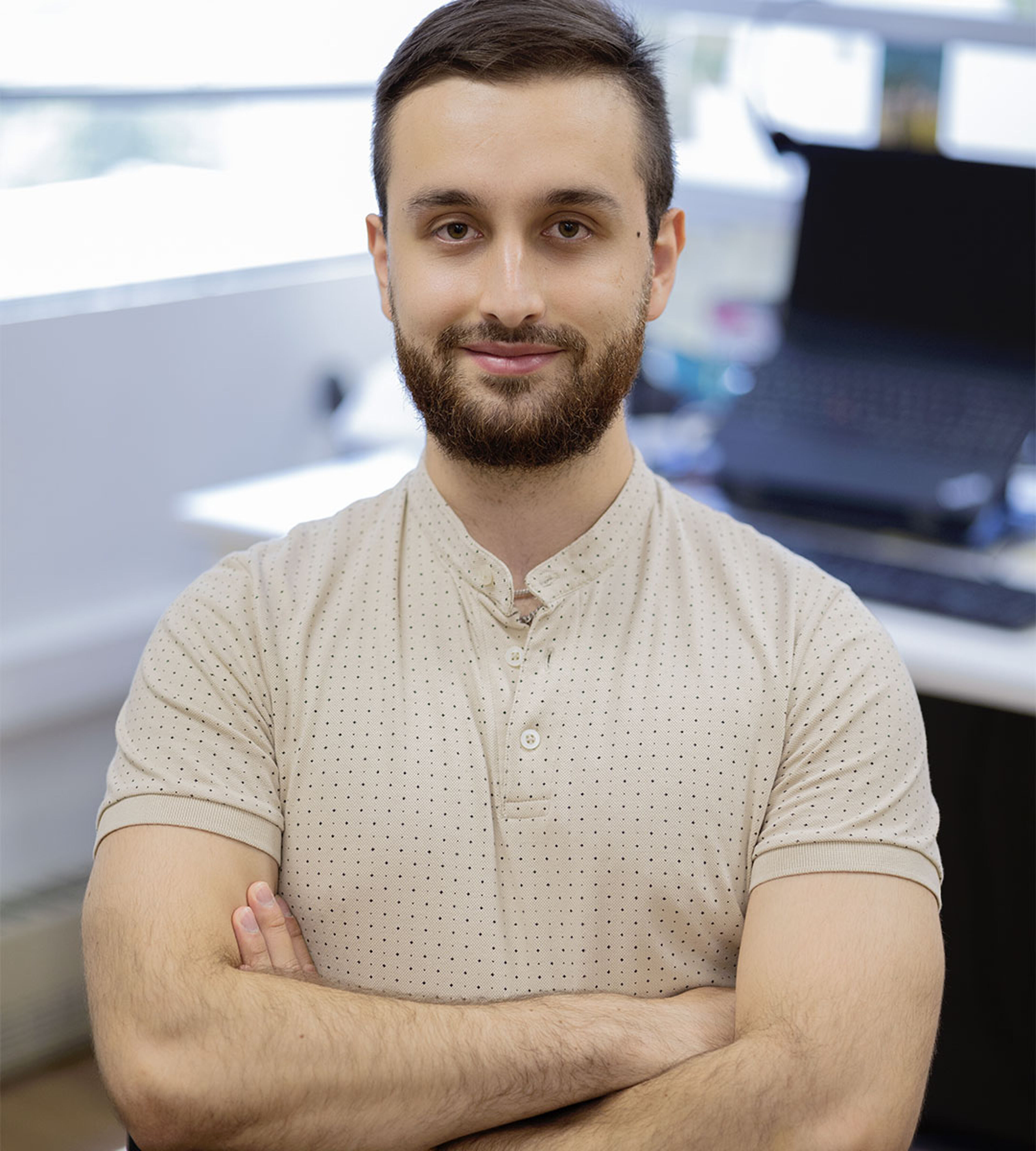 A well-groomed man with a beard in a polka-dot shirt, exuding confidence and approachability, stands in an office setting with a laptop in the background.