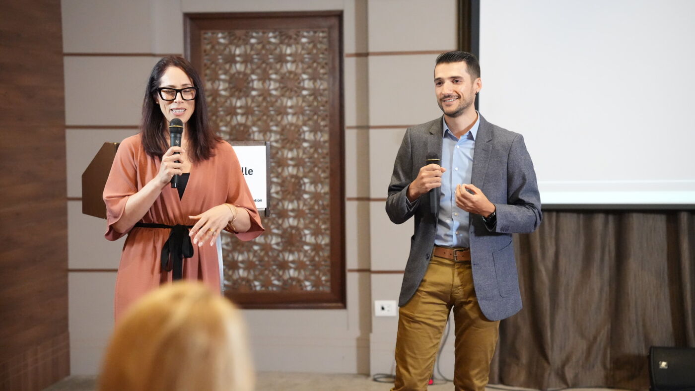 A woman and a man presenting at a business seminar, engaging with an audience off-camera.