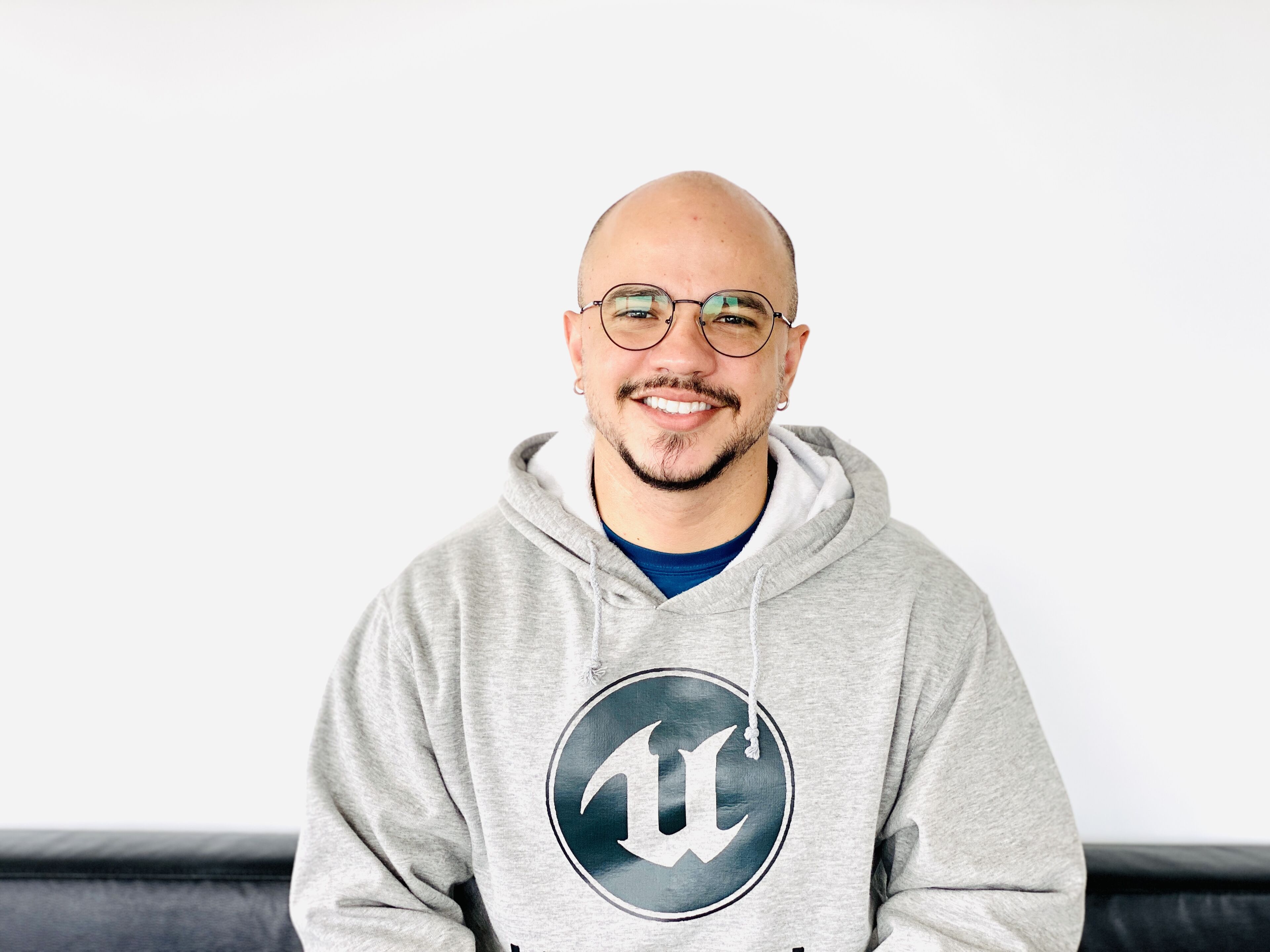 A cheerful bald man with a beard, wearing round glasses and a gray hoodie with a logo, posing against a white background.