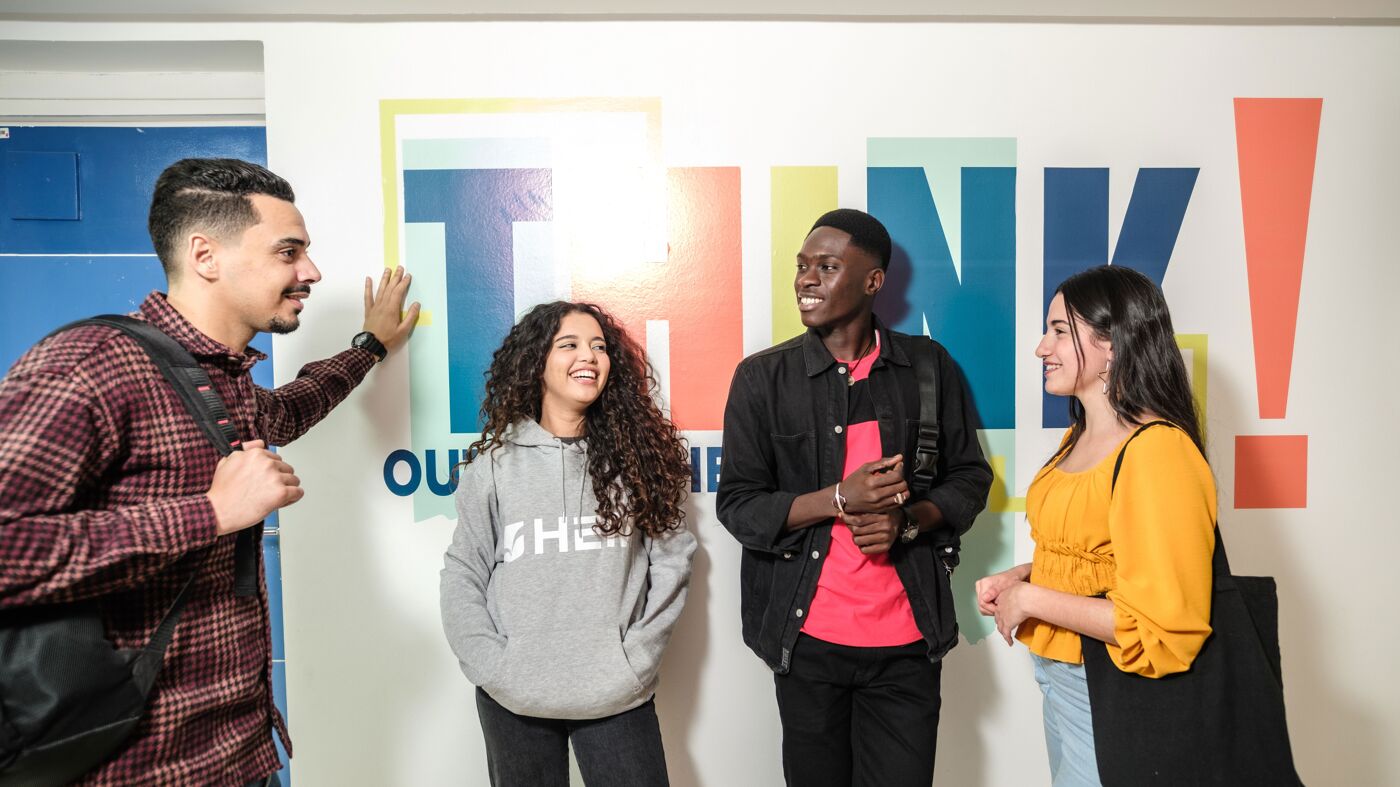 Four diverse young adults are engaging in a jovial conversation in a college hallway, with a vibrant "THINK" mural in the background.