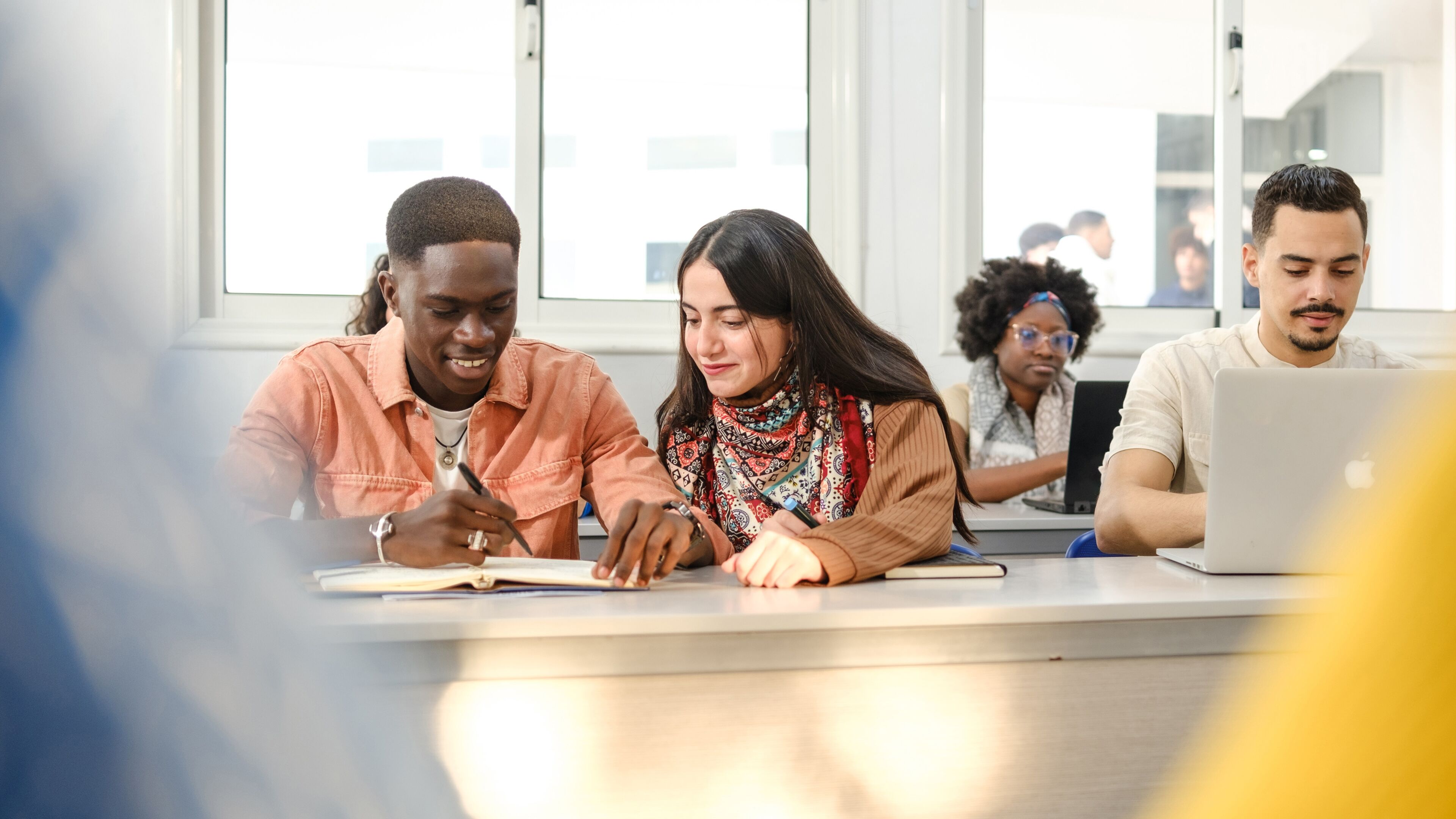 Two students collaborate on notes, with peers studying in the background.