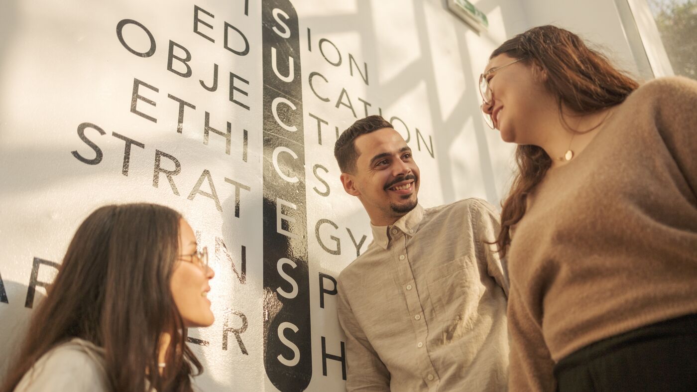 Three students converse joyfully in front of a wall with educational words.