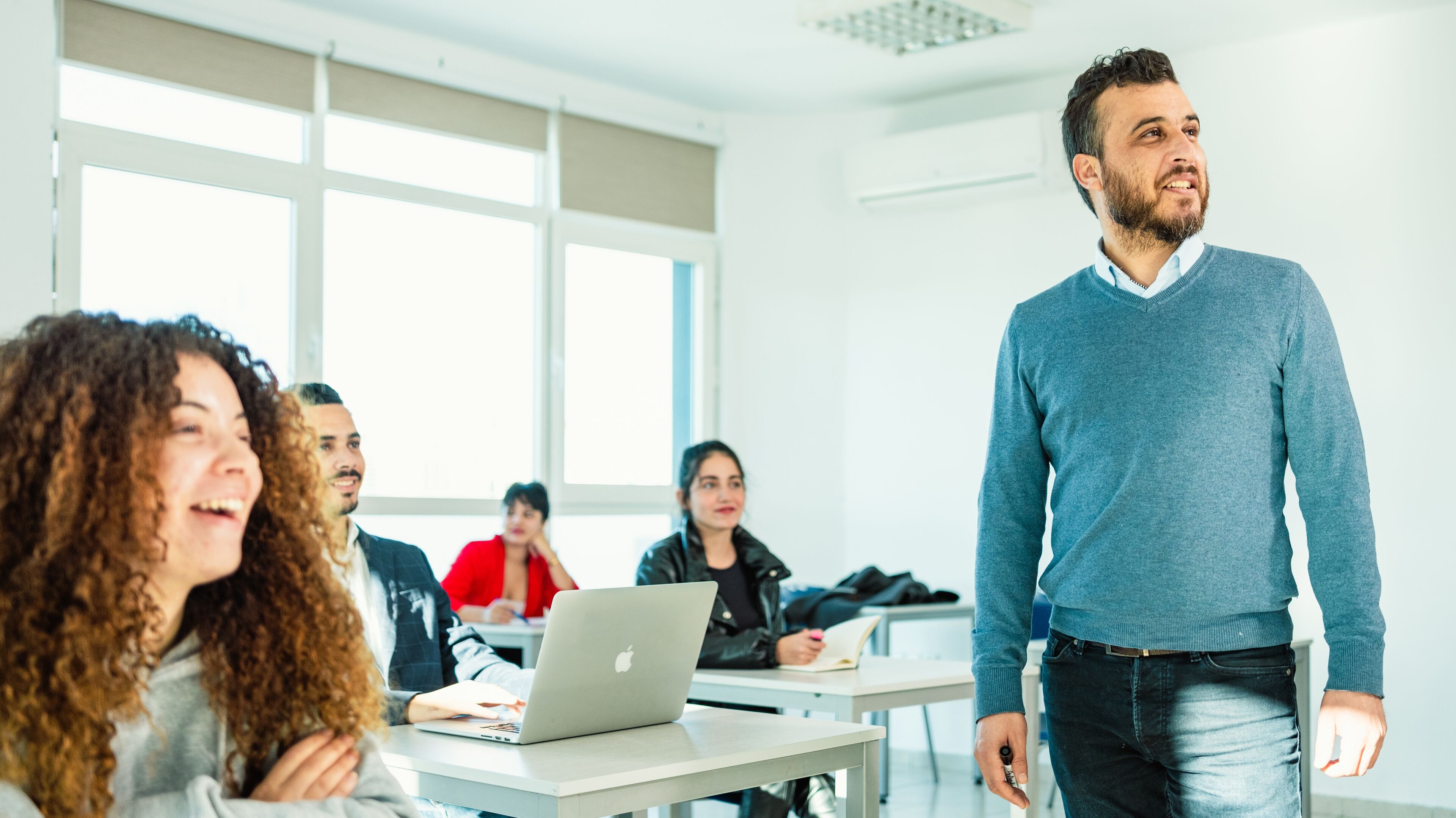 A male teacher in a blue sweater engaging with diverse students in a bright classroom, fostering an atmosphere of interactive learning.