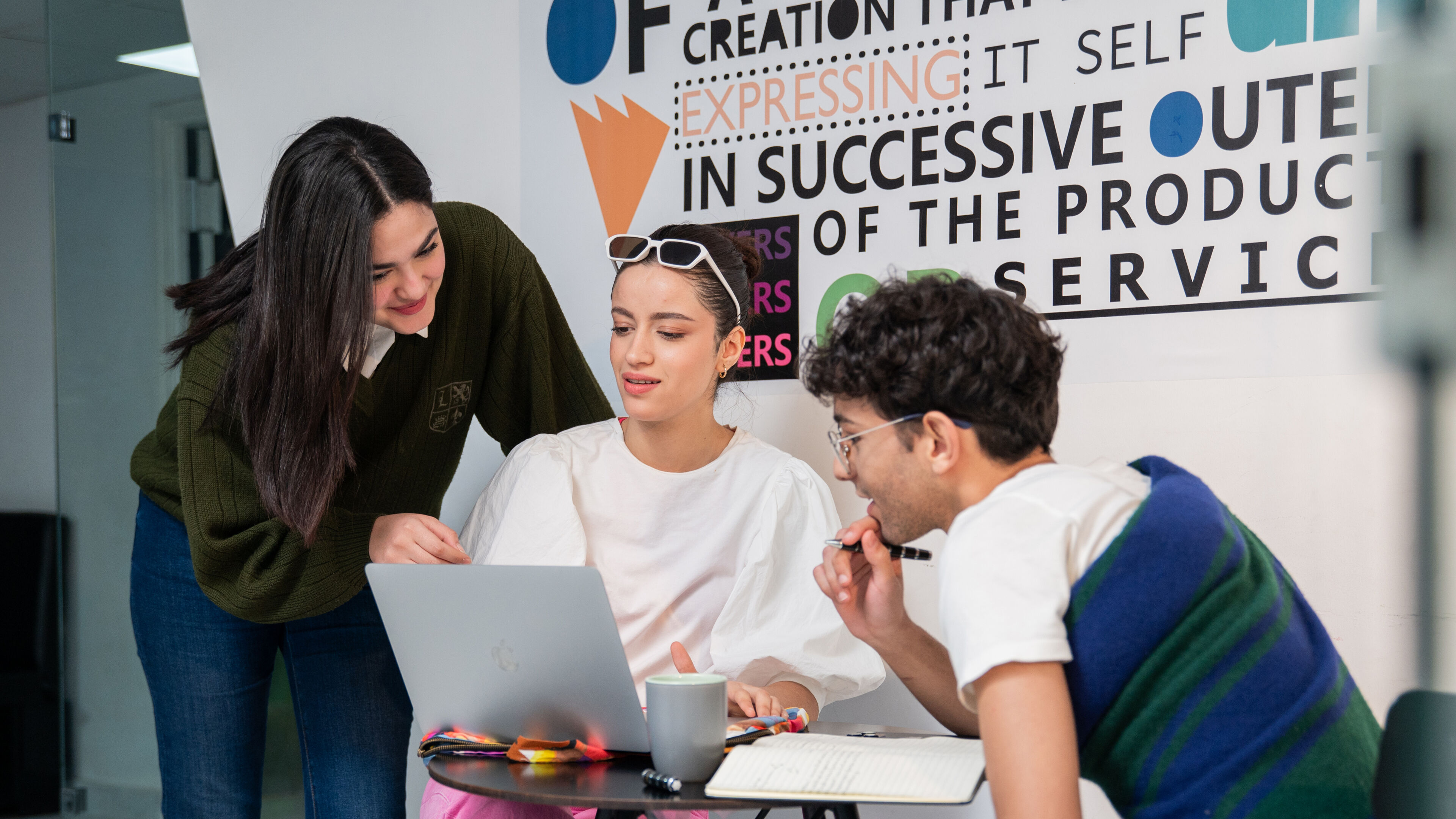 Three students discussing a project on a laptop in a creative workspace, surrounded by colorful design elements.