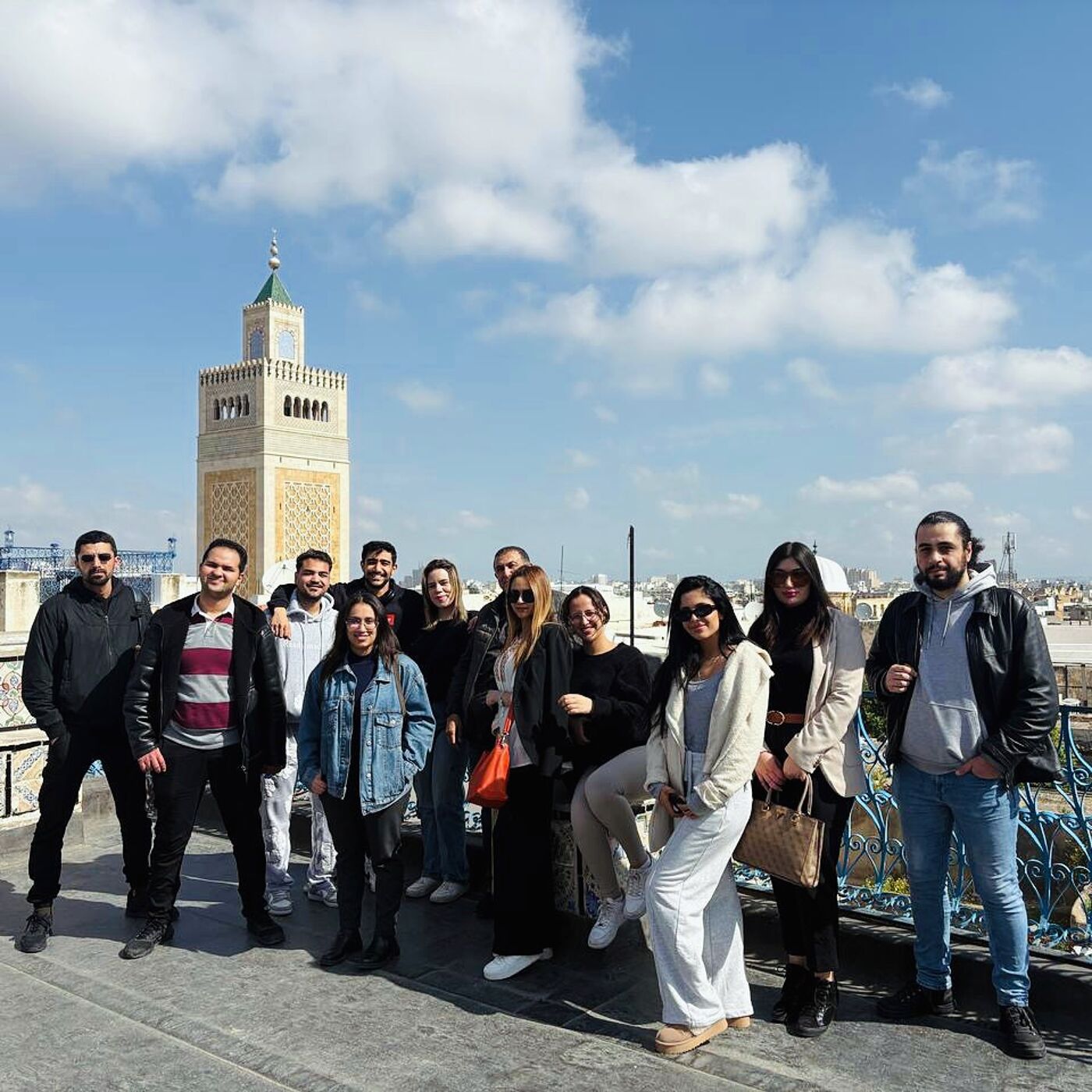 A large group of young adults stand on a rooftop, posing for a picture in front of a mosque minaret and a cityscape under a bright blue sky.