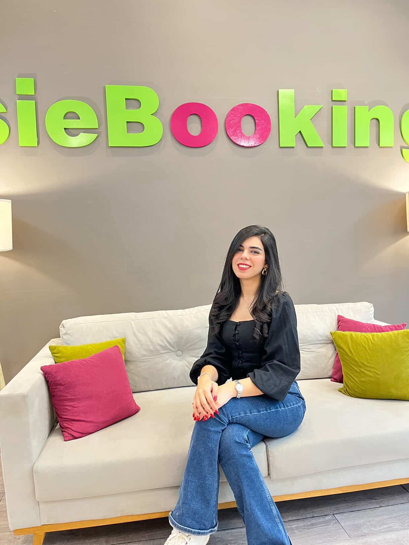 A woman with dark hair and a black top is sitting on a white couch in front of a wall with the word "sieBooking" on it. She is wearing blue jeans and has a watch on her left wrist. There are colorful pillows on the couch.