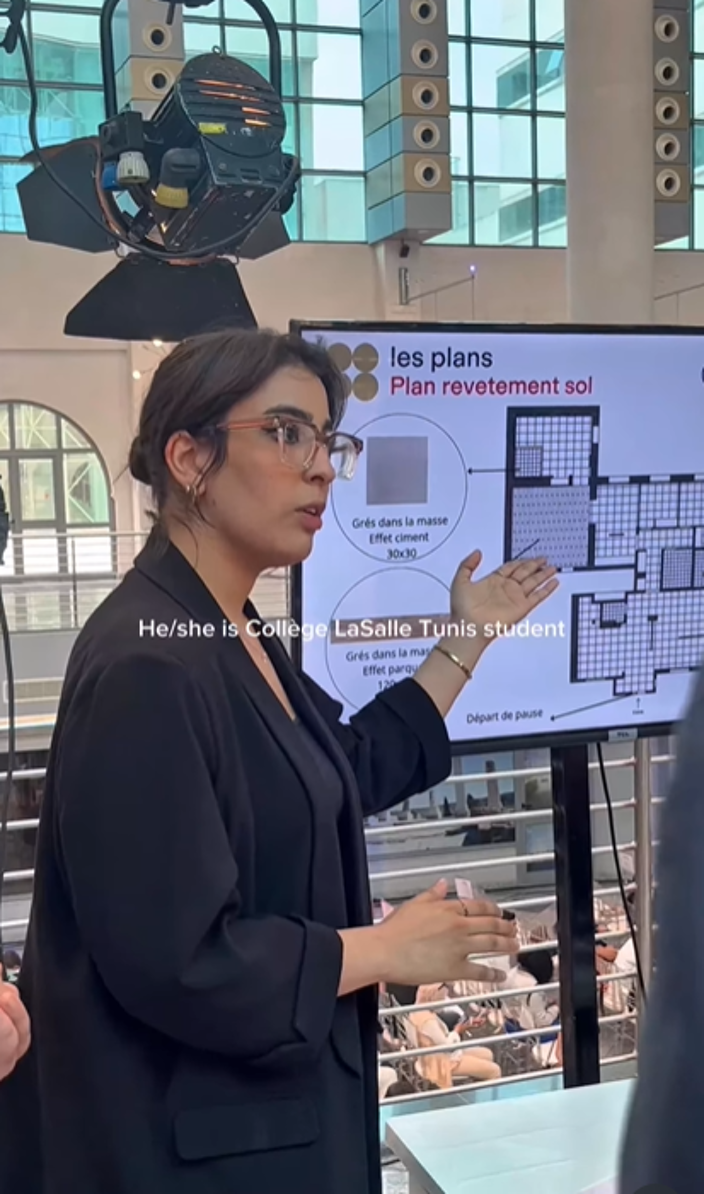 A woman in a black blazer and glasses is presenting a floor plan on a screen. The screen displays architectural details and French text. She appears to be a student at College LaSalle Tunis.