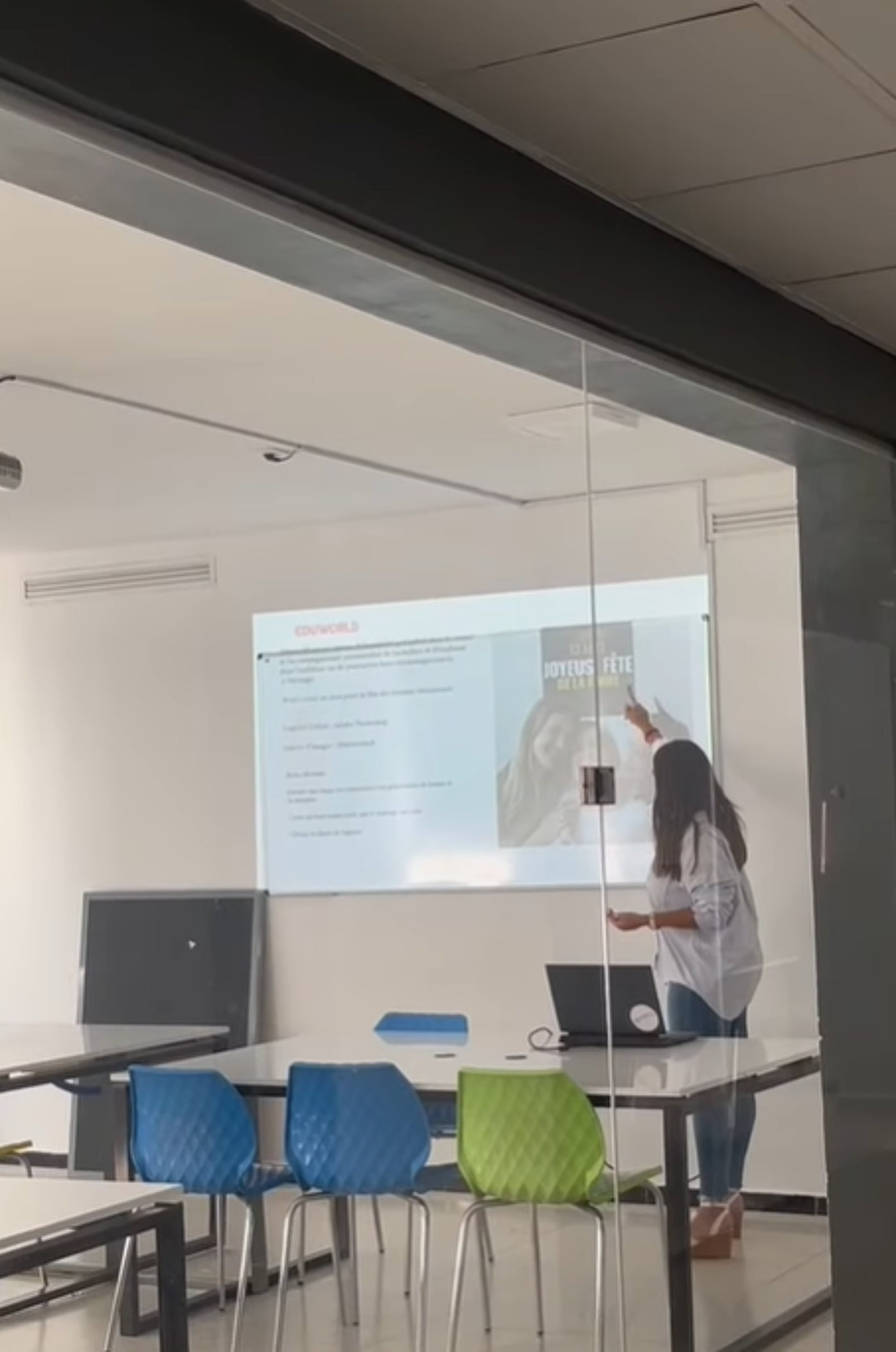 A woman is giving a presentation in a classroom. She is pointing to a slide on a projector screen that features a book cover. The room has tables, chairs, and a blackboard.