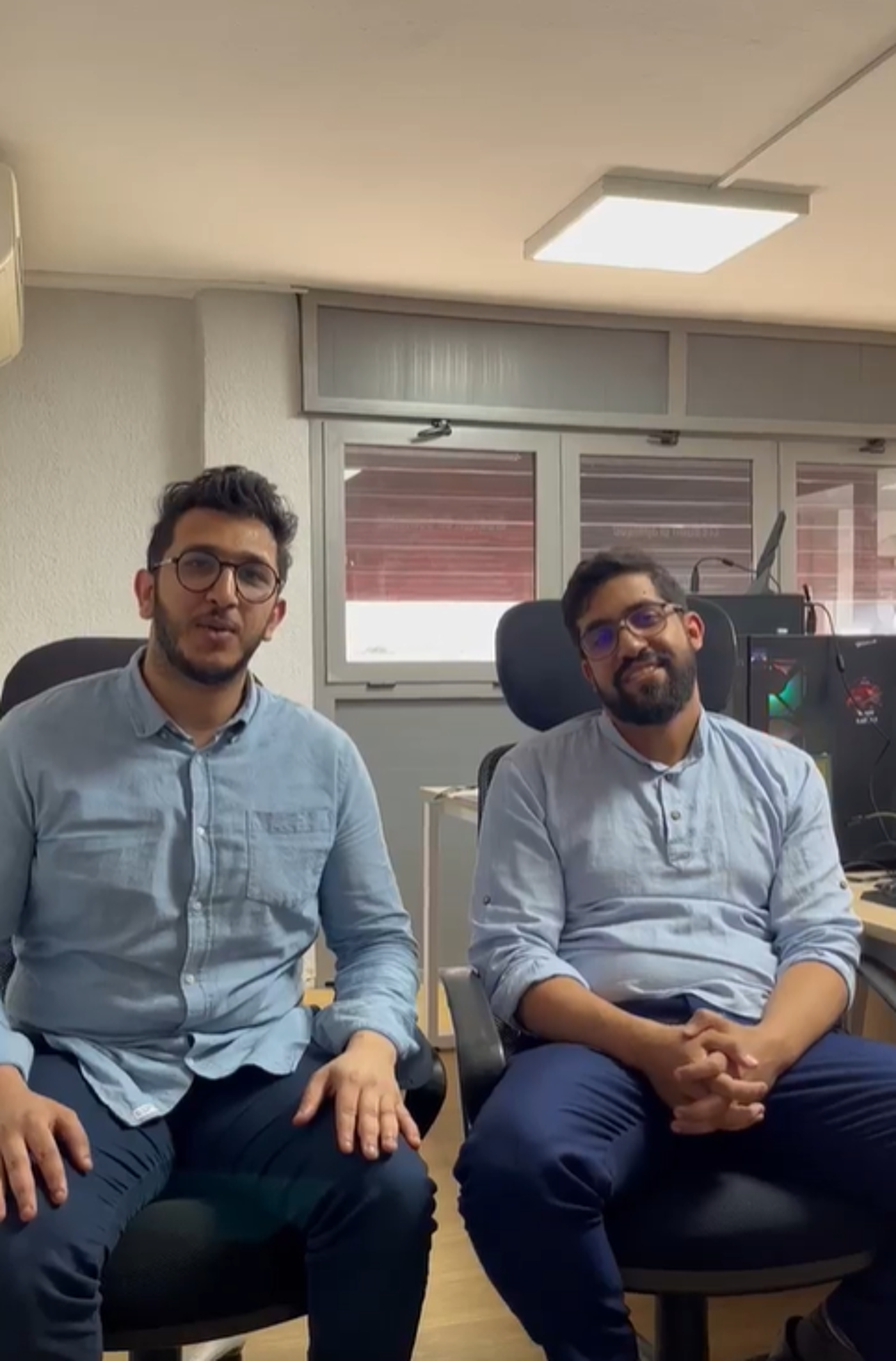 Two young men with beards and glasses are sitting in office chairs, wearing blue shirts and dark pants. They appear to be in an office setting with computers and a window visible in the background.