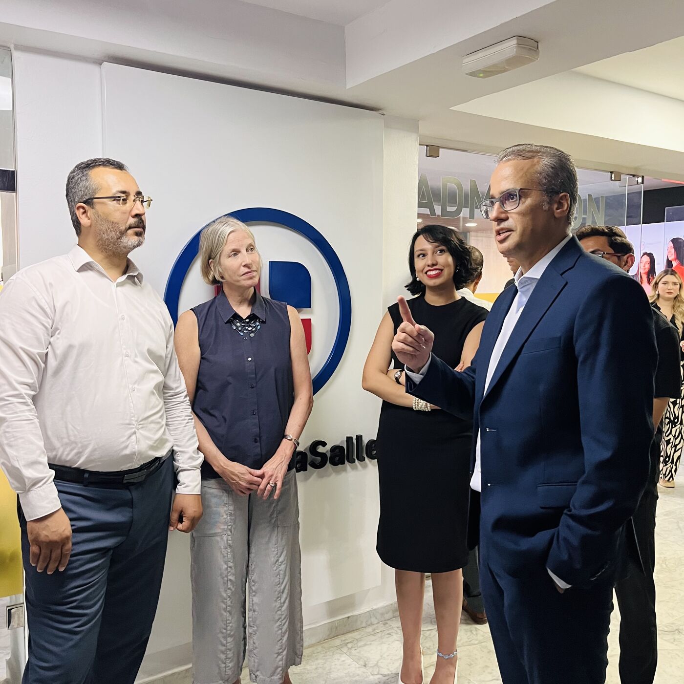 A group of five people are standing in front of a La Salle logo. A man in a suit is speaking while gesturing with his right hand. The others are listening attentively. The setting appears to be an indoor event or gathering.