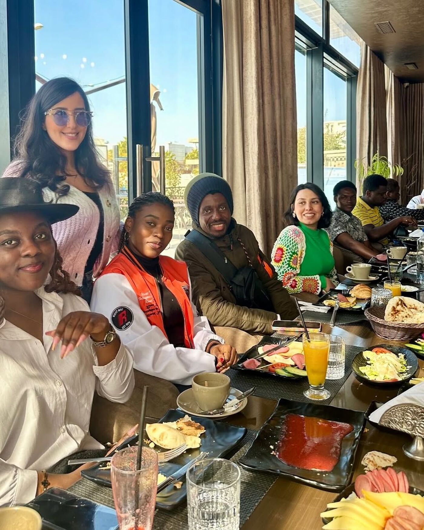 A group of diverse people are seated around a table laden with food and drinks at a restaurant, enjoying a meal together.