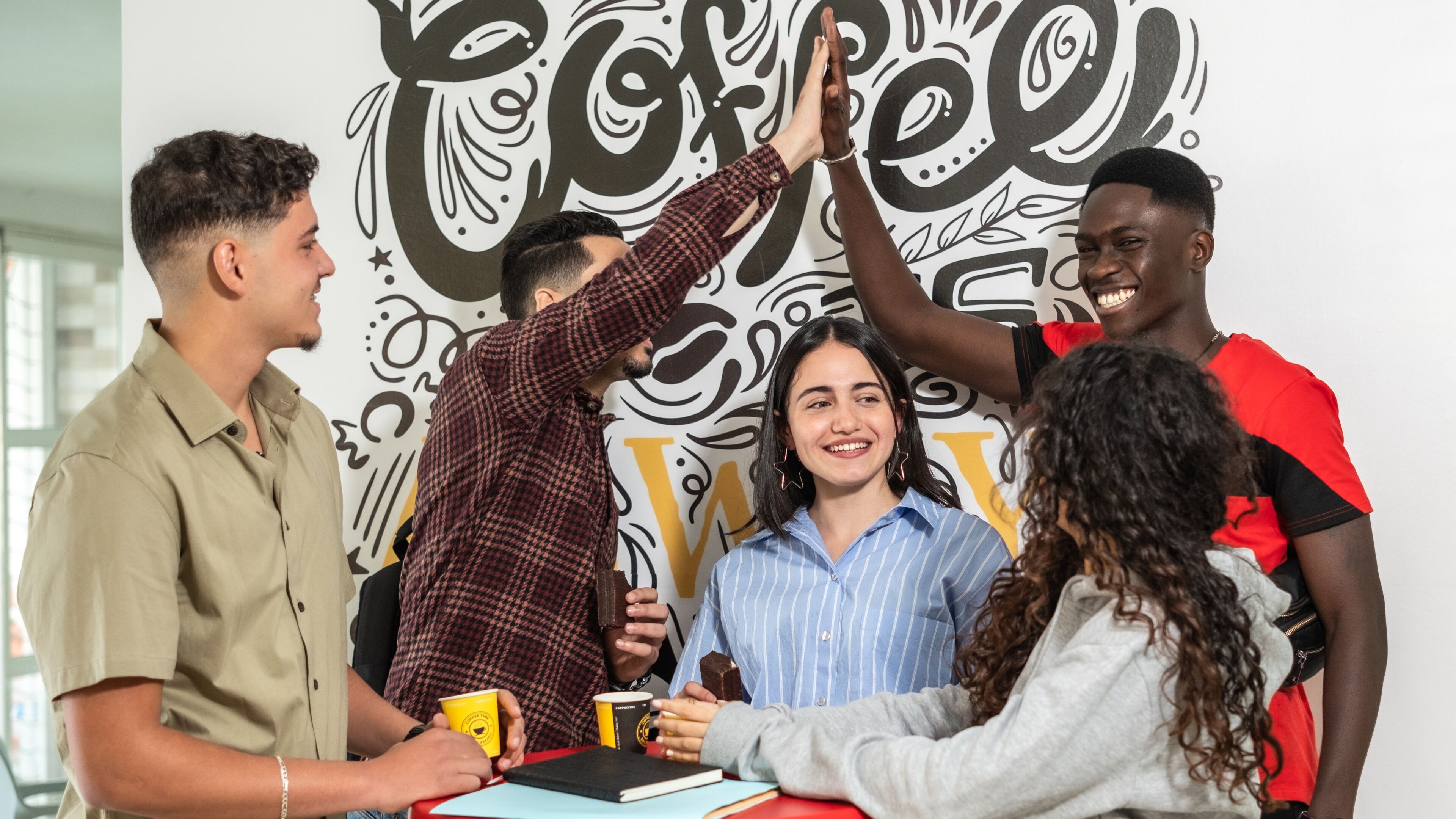 A diverse group of students joyfully giving each other a high five in a moment of celebration, with a creative mural in the background.