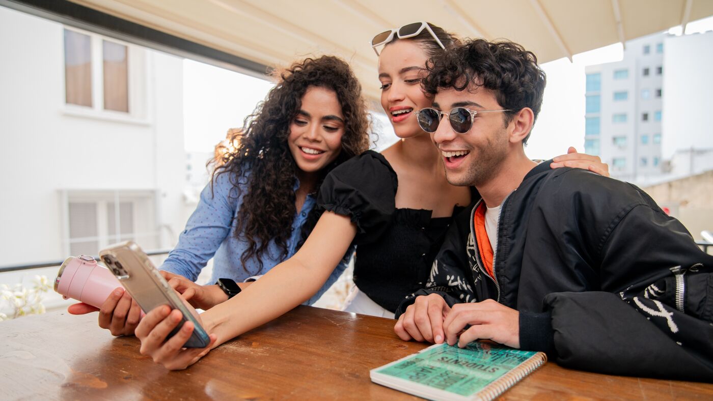 Three friends enjoy a fun moment, sharing content on a smartphone together, outdoors during the day.