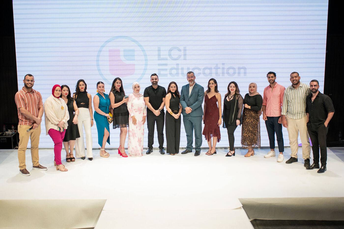 A group of people are posing for a photo on a stage with the LCI Education logo in the background. They are dressed in various outfits.