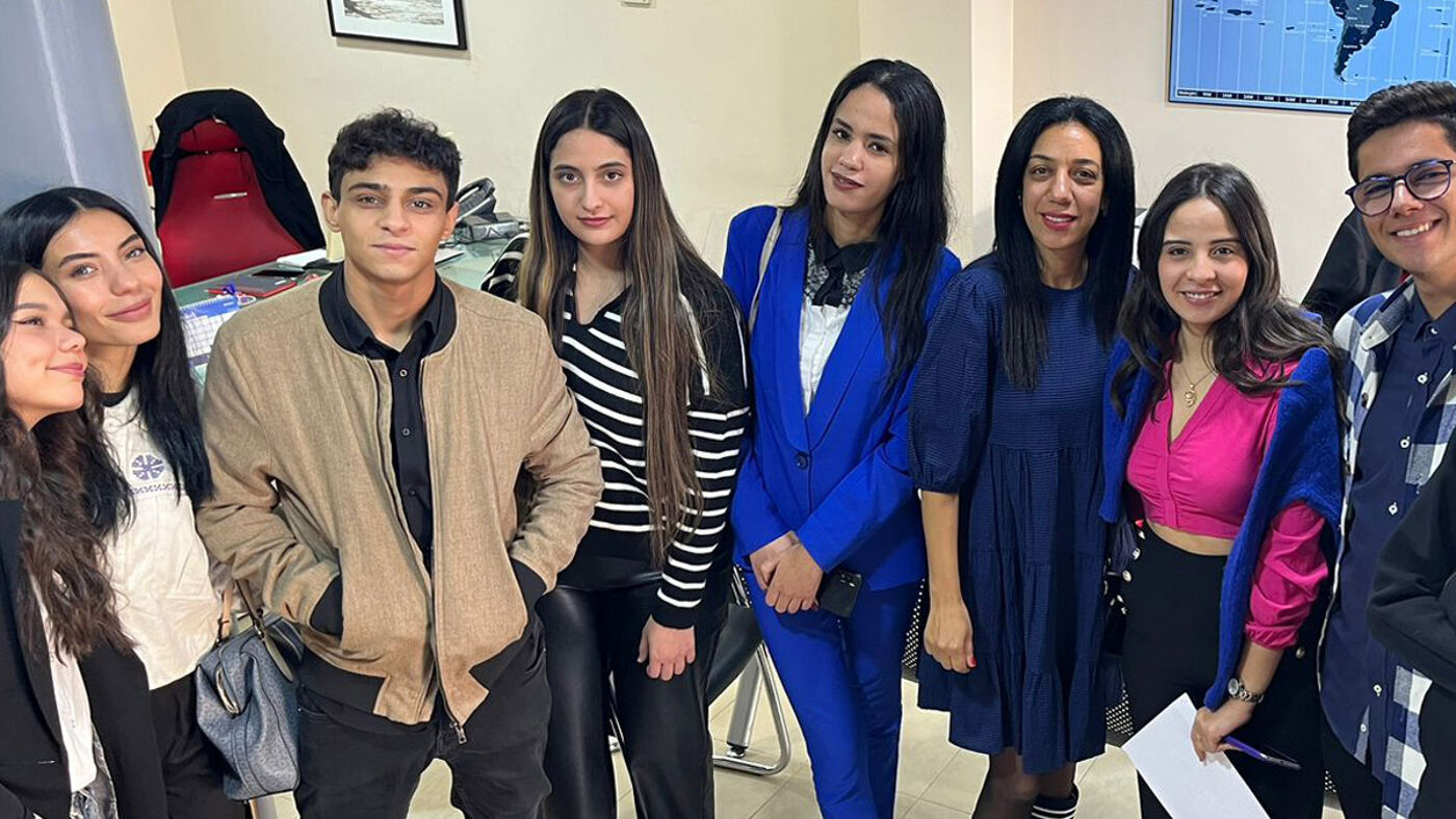 A diverse group of eight individuals, dressed in business casual attire, smiling for a group photo in an office setting.