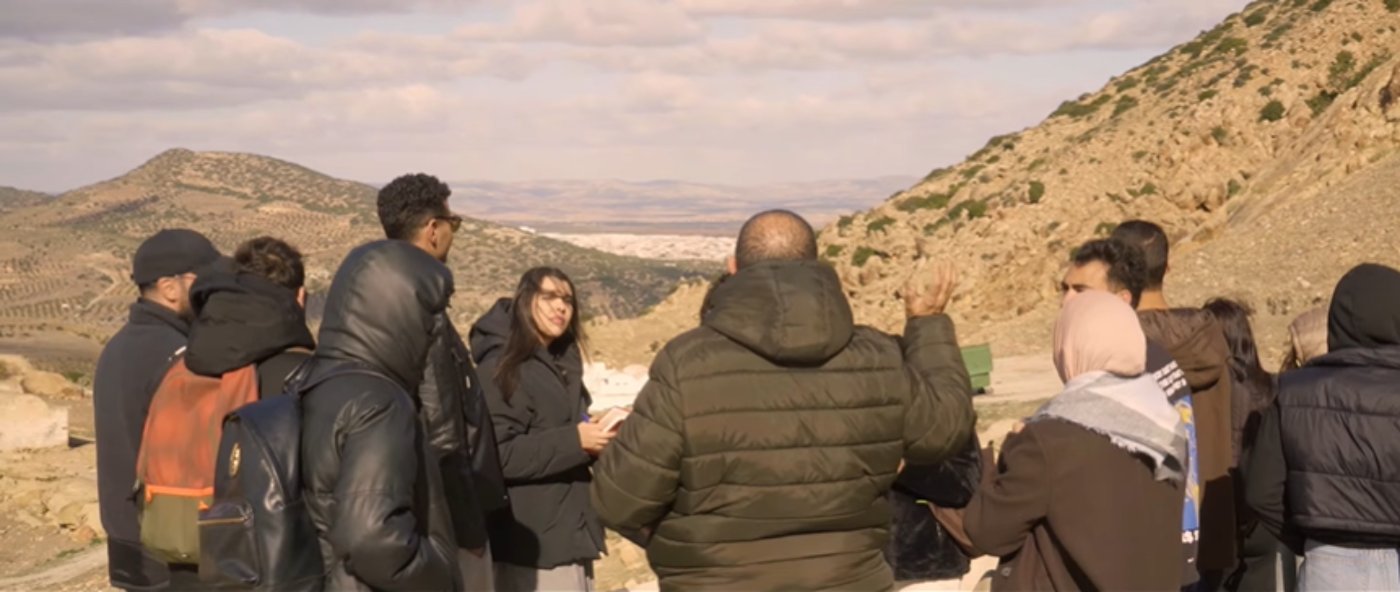 A group of people are gathered outdoors, possibly on a hillside, with a scenic view of mountains and a distant town. They seem to be listening to someone.