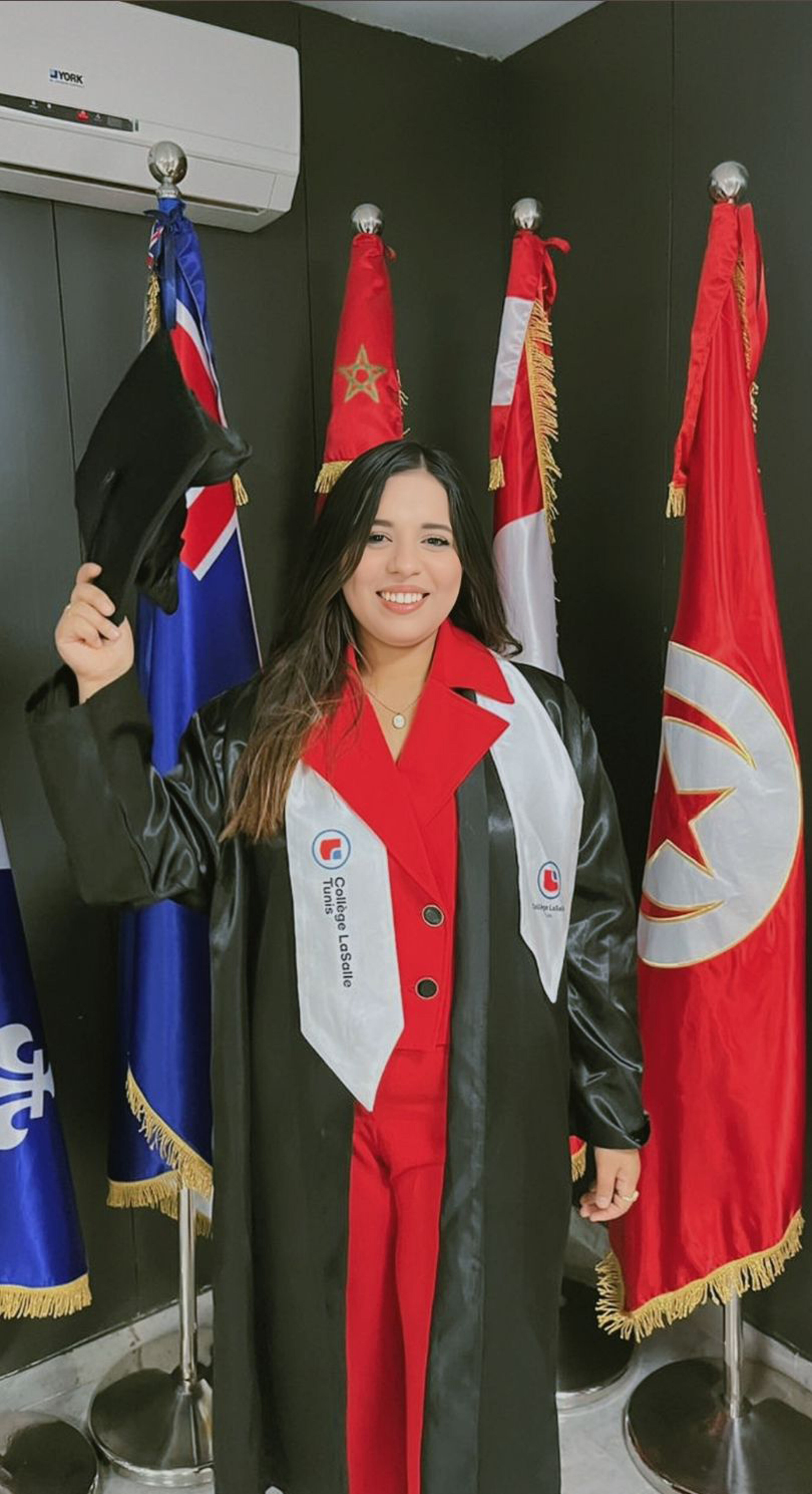  A smiling graduate poses in cap and gown at Collège LaSalle Tunis, holding her cap proudly. She stands in front of several international flags, symbolizing the global community and accomplishment.