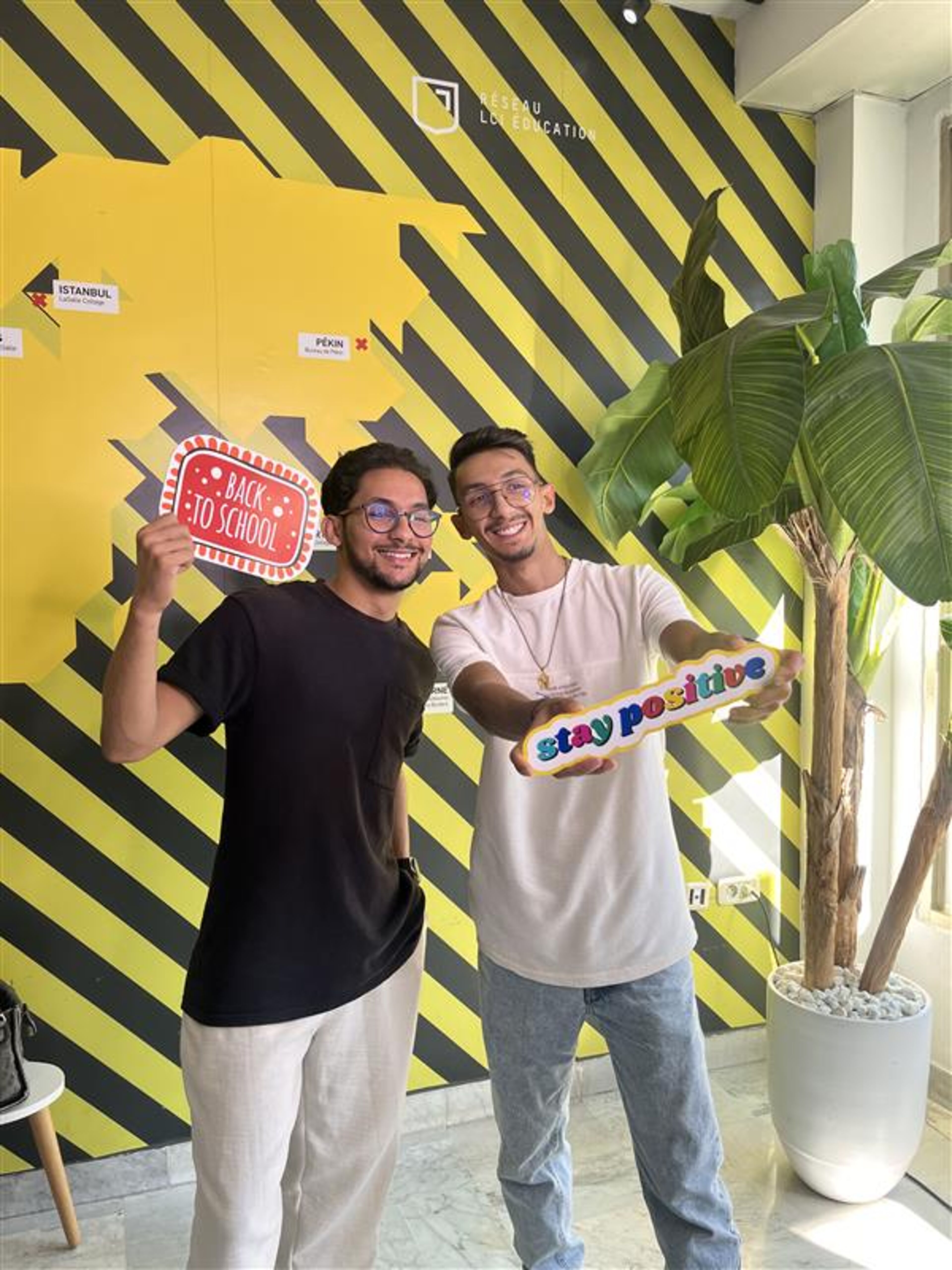 Two young men smile and hold "Back to School" and "Stay Positive" signs in a vibrant setting with a yellow and black background. The playful atmosphere and encouraging messages reflect a cheerful back-to-school vibe.