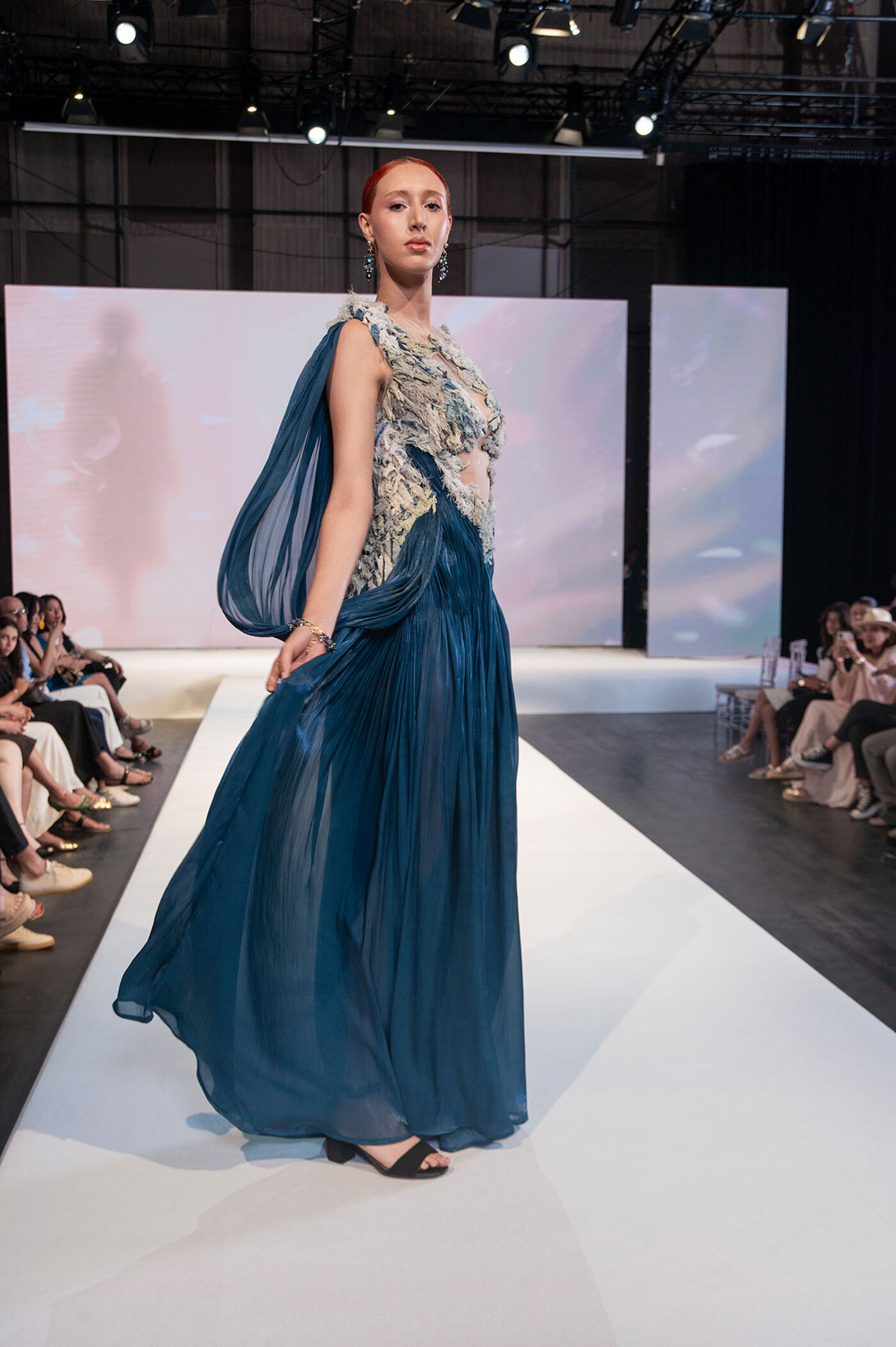 A woman is walking on a runway wearing a long, flowing blue dress with a patterned bodice. The dress has a cape-like sleeve and she is wearing black sandals. There are people seated on either side of the runway.