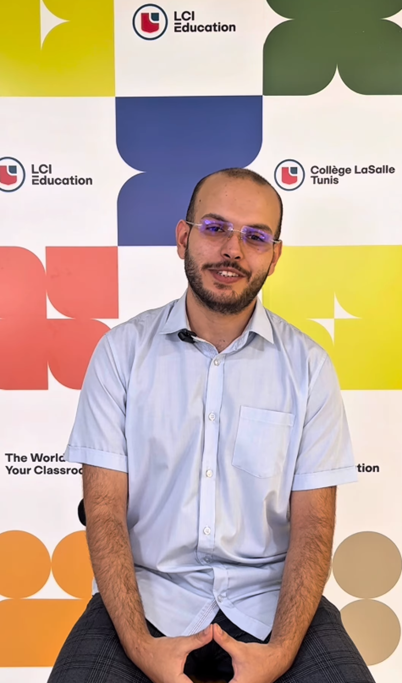 A man with glasses and a beard is sitting in front of a colorful backdrop featuring logos of LCI Education and Collège LaSalle Tunis. He is wearing a light blue shirt and has his hands clasped together.