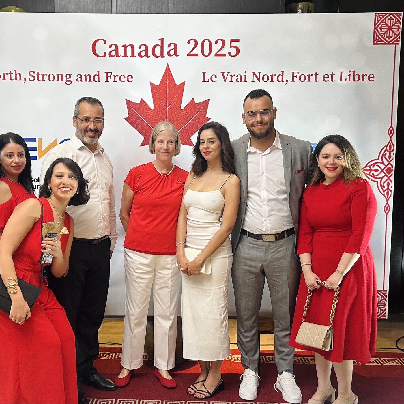 The image shows a group of seven people standing in front of a backdrop that reads "Canada 2025" and "Le Vrai Nord, Fort et Libre". Most of the people are wearing red, and they appear to be at an event or gathering.