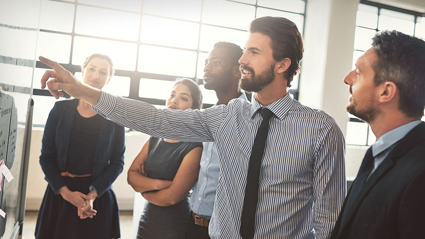 A man with a beard leading a presentation, pointing at a whiteboard, with attentive colleagues around him.