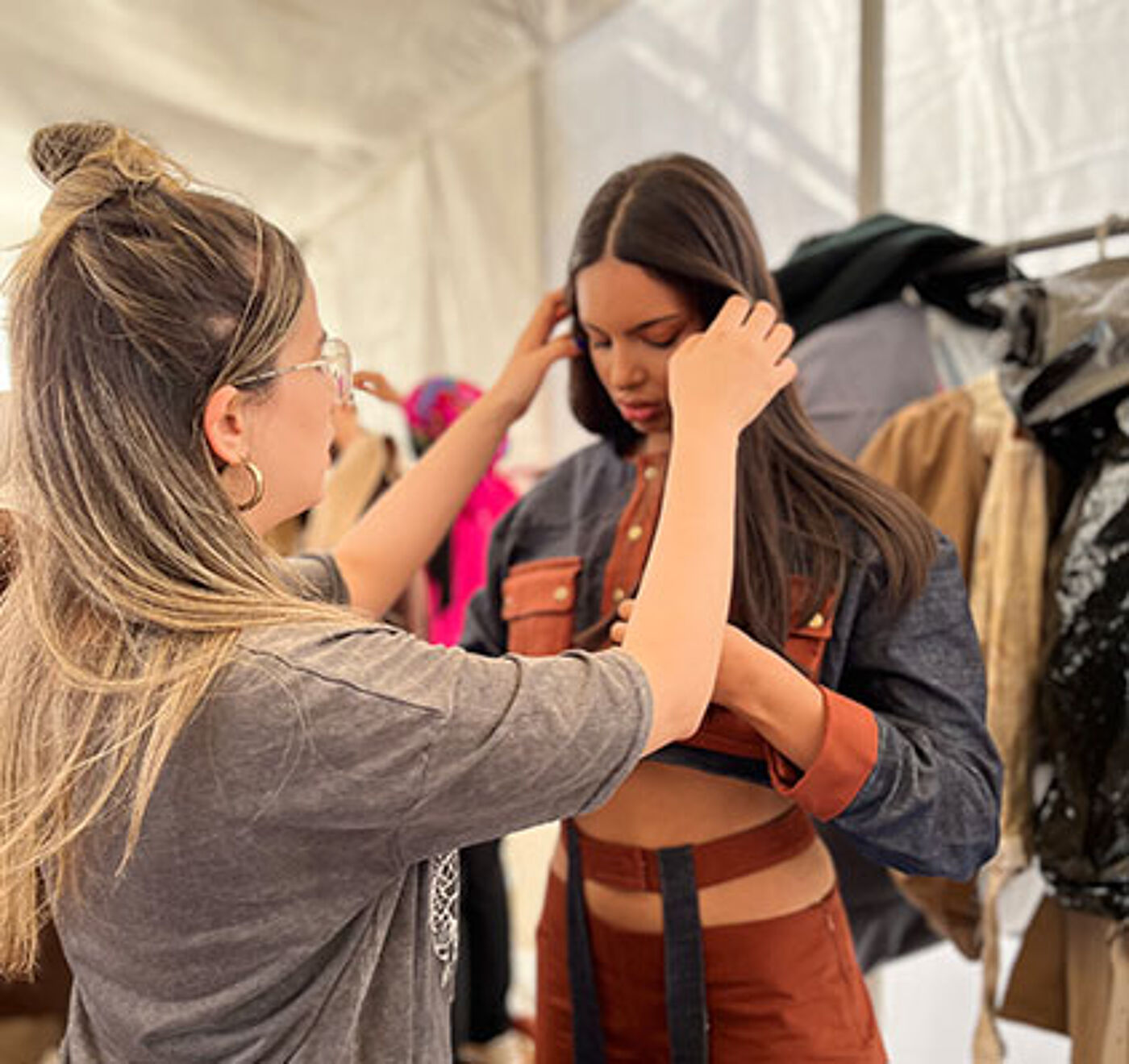 A focused stylist secures the fit of a leather outfit on a model within the busy environment of a fashion show tent.