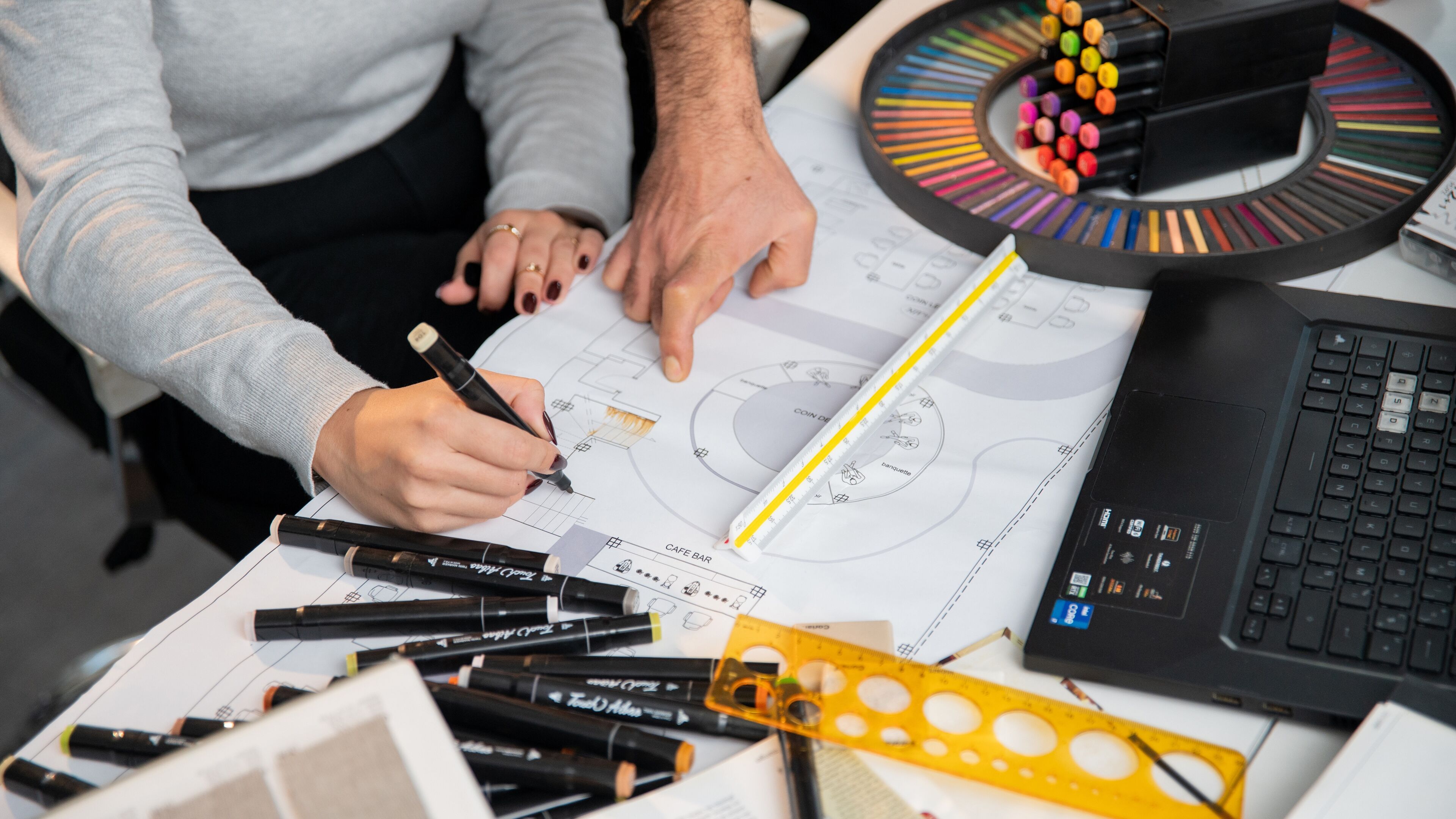 Deux individus collaborent sur un plan architectural, entourés d'outils de conception et de palettes de couleurs.