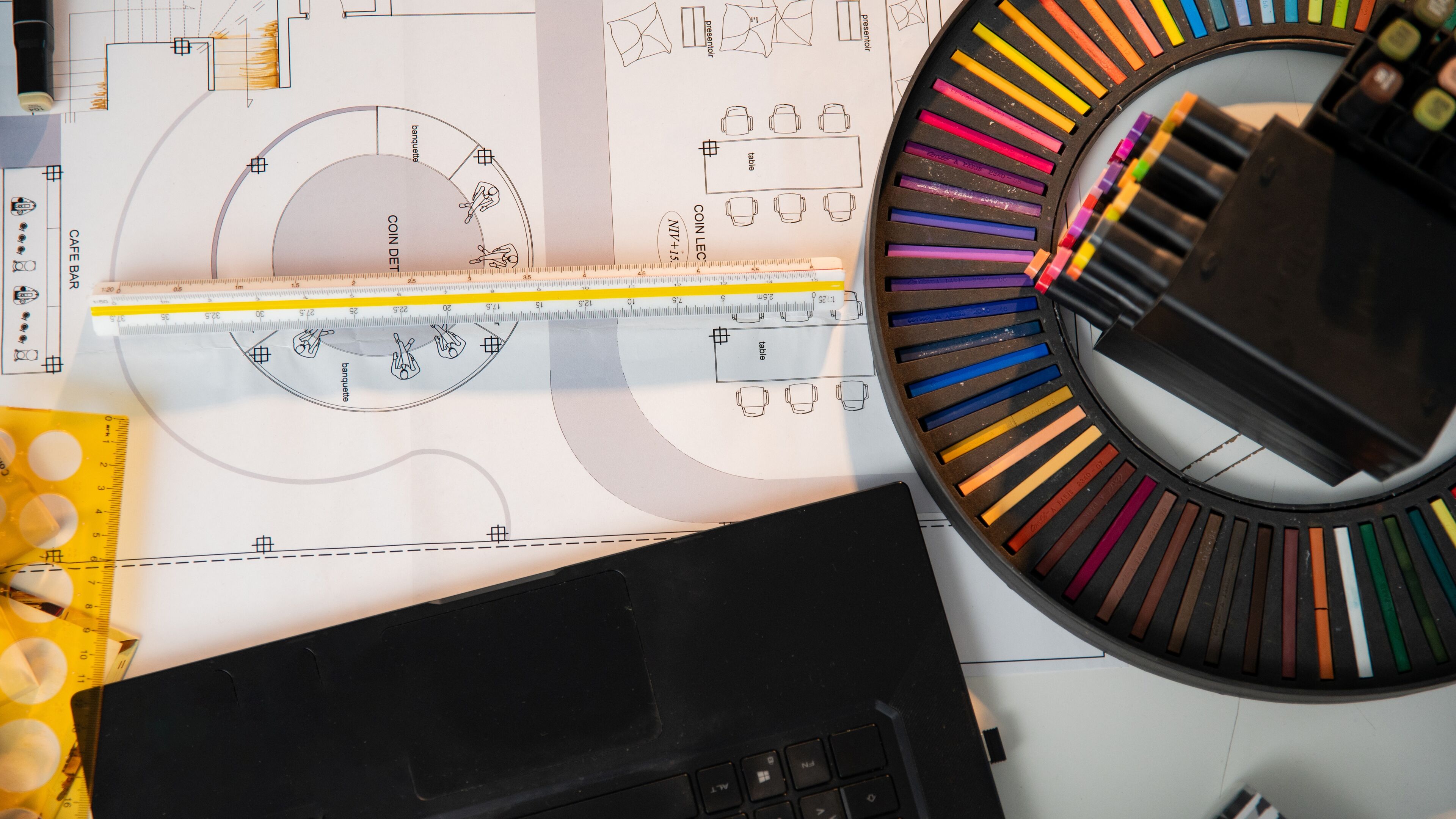 Vue rapprochée d'un équipement de dessin technique professionnel, comprenant une règle, un rapporteur et des plans architecturaux, à côté d'un étui circulaire de crayons de couleur éclatants.