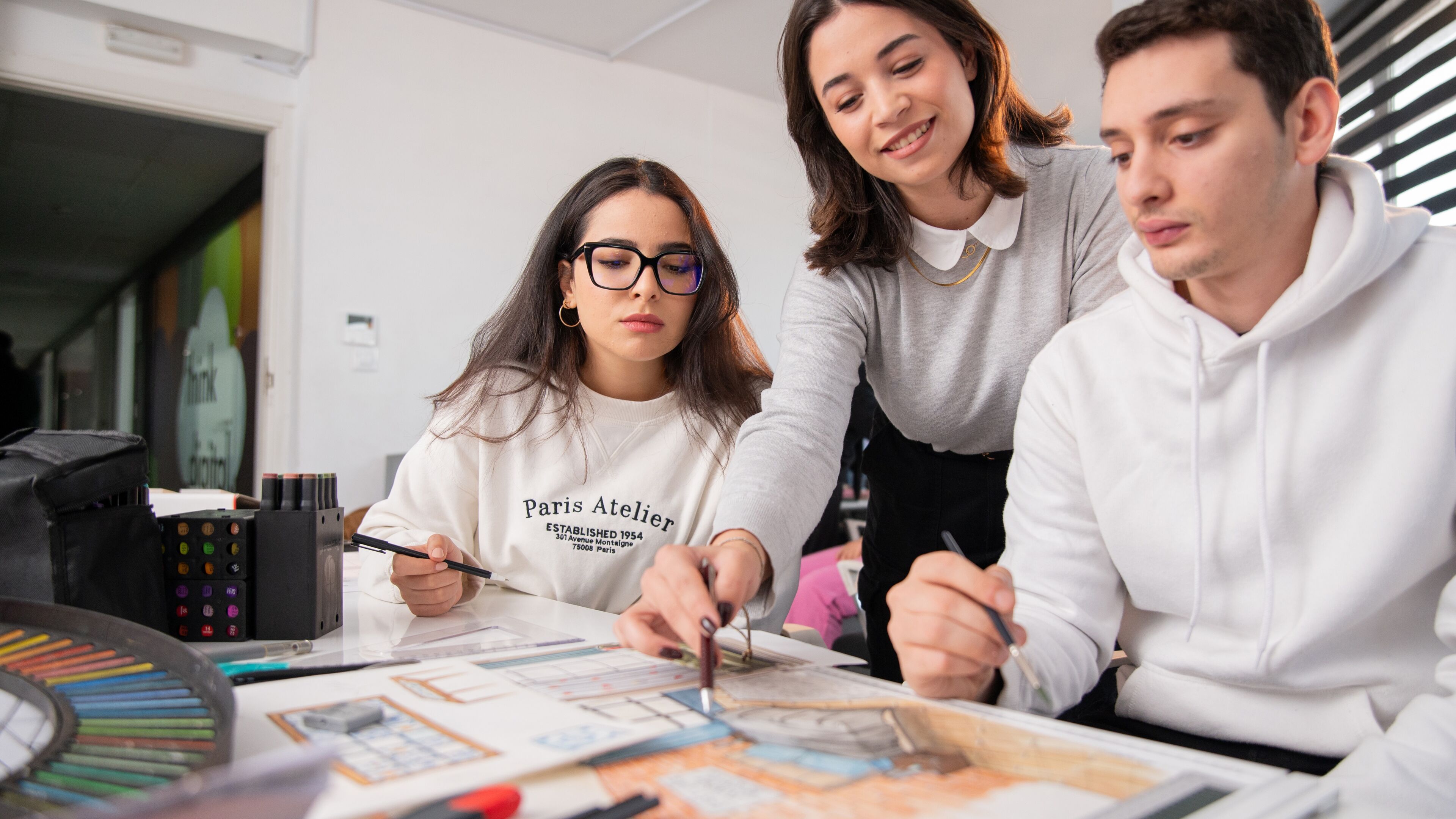 Three art students engage in a collaborative project, with color palettes and markers on the table.