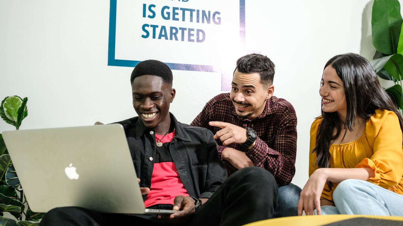 Three colleagues share a light moment around a laptop in an informal office setting.