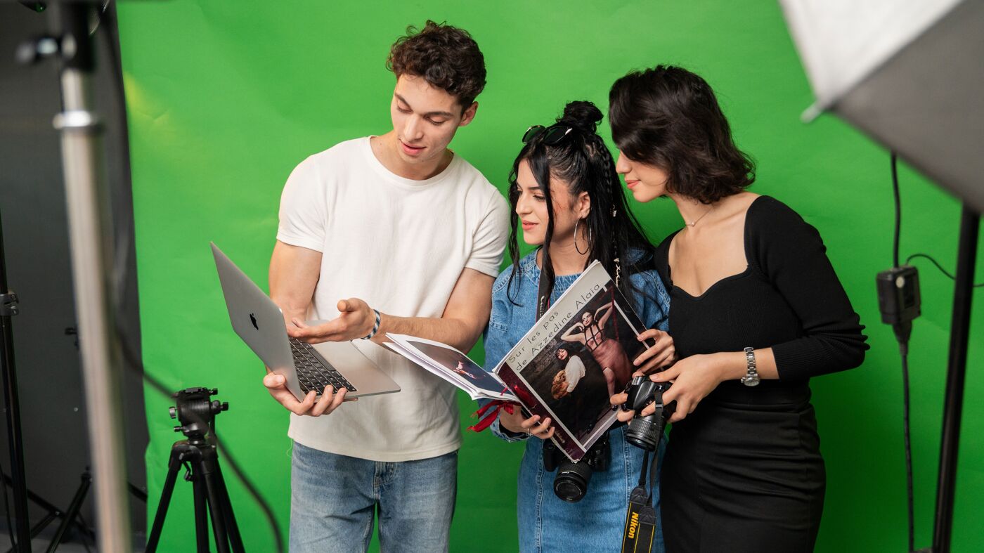 Three individuals collaborate behind the scenes, with a laptop and camera equipment, against a chroma key backdrop.